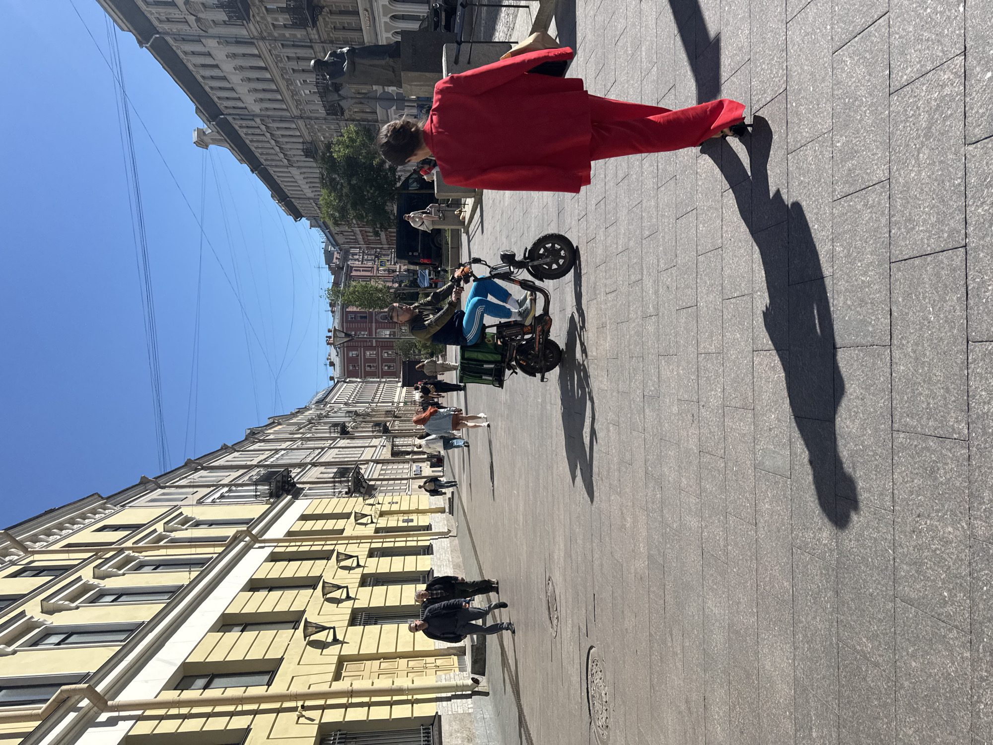 A person in a bright red suit walks along a sunlit cobblestone street lined with tall, ornate buildings. The street slopes downwards, with a clear blue sky overhead, suggesting a pleasant day. The person in red is the dominant subject, casting a long shadow on the pavement. They appear to be walking away from the camera. In the mid-ground, a person is riding a small electric scooter, also casting a shadow. Other people are visible further down the street, some walking, others standing. The buildings on the left are a pale yellow with white trim and dark windows, typical of St. Petersburg architecture. Overhead, several power lines crisscross the sky. The overall scene depicts a moment of urban life on a bright, sunny day. The time of day appears to be late morning or early afternoon, given the length and angle of the shadows. There is no visible text in the image.