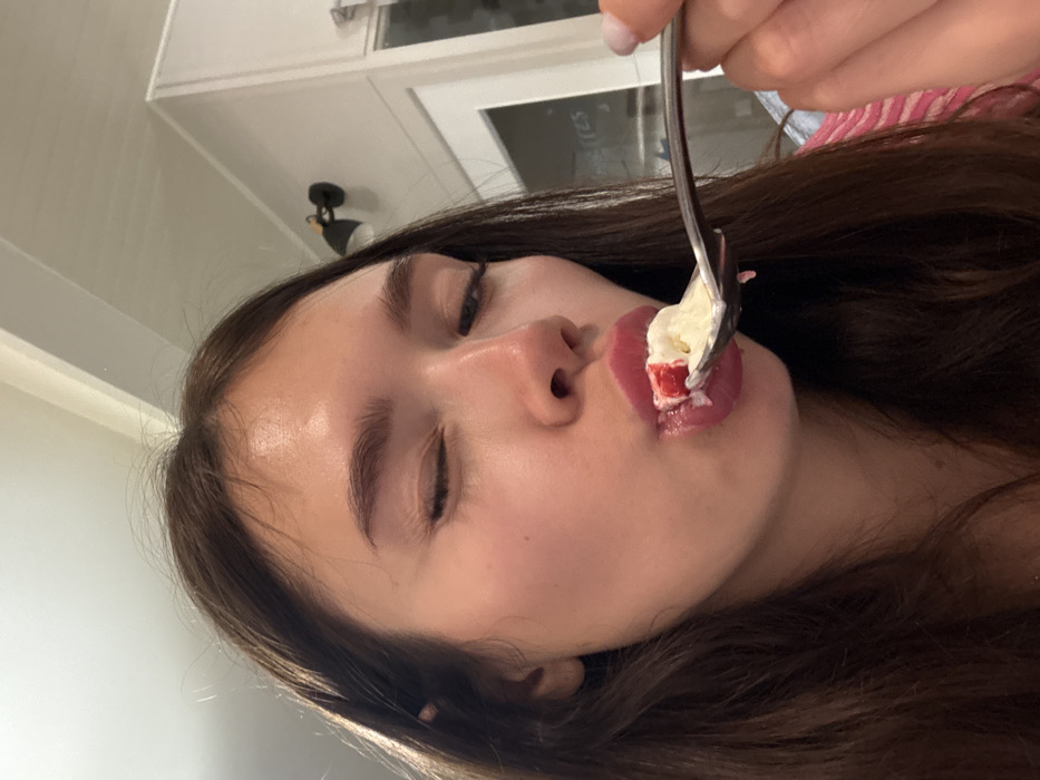 A woman lies on her side, eyes closed, eating a bite of white cake with a red topping from a fork. Her long, dark brown hair falls around her face. She has fair skin and full lips. The background reveals a white wall and a glimpse of white kitchen cabinets. A black, modern light fixture is visible above her head. The image appears to be taken indoors with natural lighting.