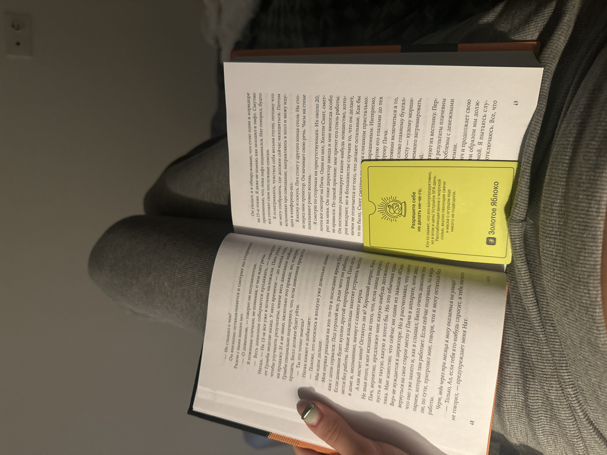 A person is reading a book indoors, resting the open book on their lap. The book is a paperback with a white cover and black text. A yellow card is placed on the open pages. The scene appears to be in a home or similar private setting. No specific location in Moscow, Russia, can be determined from the image.