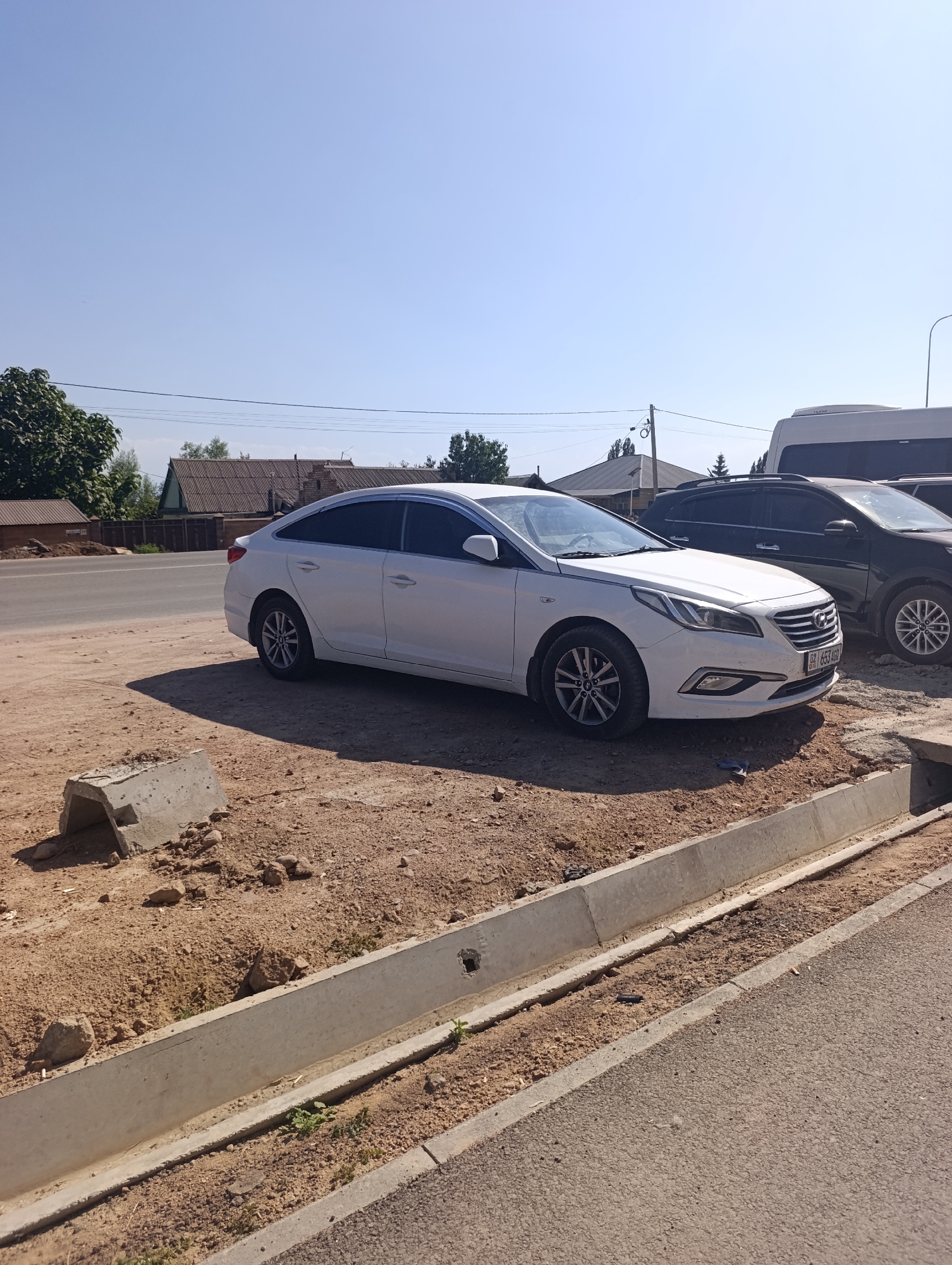 In Bosteri, Kyrgyzstan, a white sedan is parked on a dirt shoulder next to a paved road. The car is positioned facing right, with its front end slightly obscured by a dark SUV parked behind it. A white minivan is also visible in the background, parked to the right of the dark SUV. The environment is dry and dusty, with several concrete structures and debris scattered on the ground near the car. Residential buildings with tiled roofs and trees are present in the background, suggesting a semi-urban or rural setting. The sky is clear and blue, indicating a sunny day.