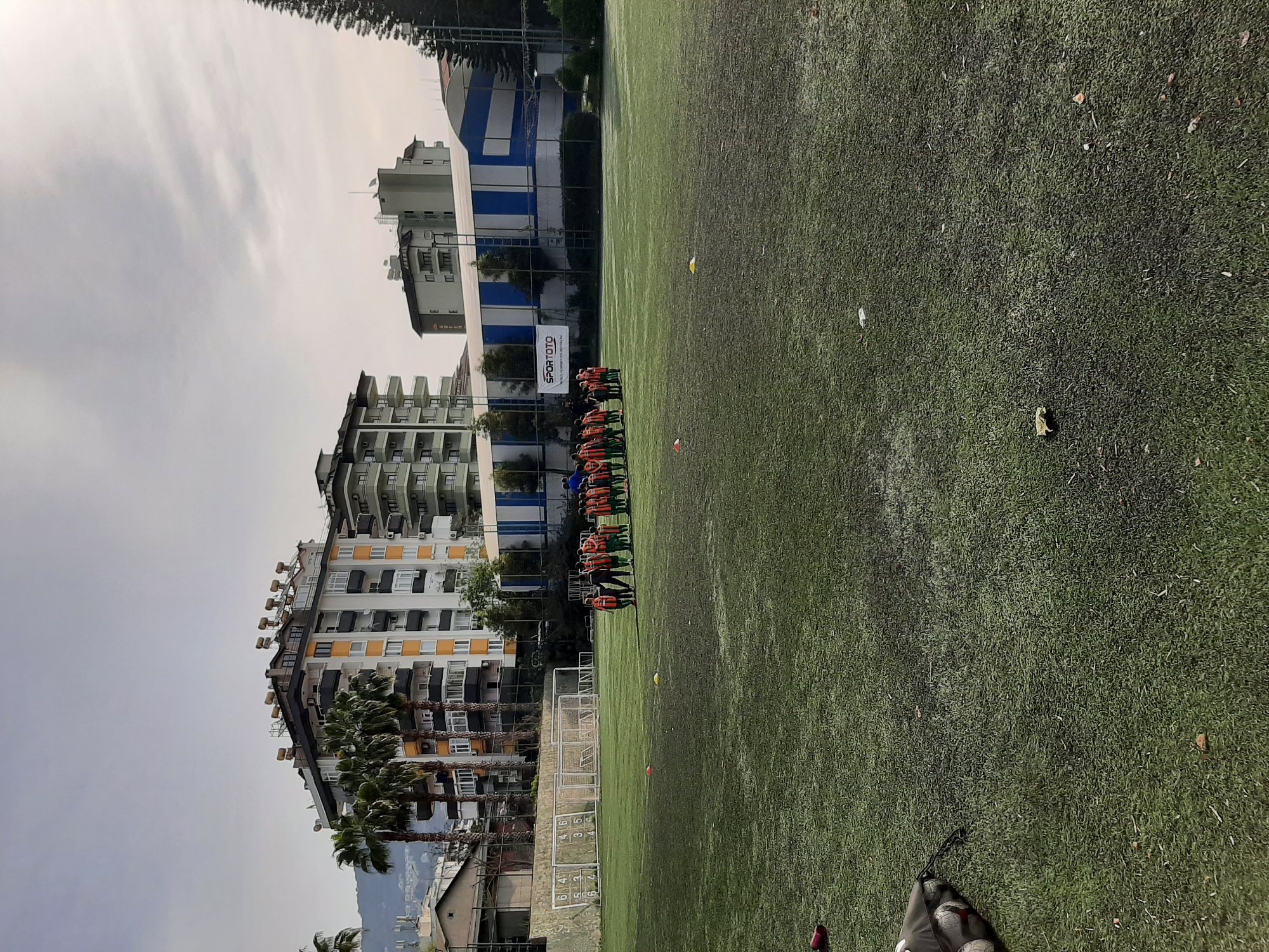A slightly tilted outdoor medium shot depicts a soccer field in the city of Alanya, Turkey. The field is green with scattered small yellow and orange cones. A group of people stand in a line near the goal, likely a soccer team. In the background, there are buildings of various heights and colors, along with palm trees and other vegetation. The sky is cloudy. At the bottom of the image, a partial view of a soccer ball bag is visible. A white sign with “SPORTOTO” is behind the group of people.