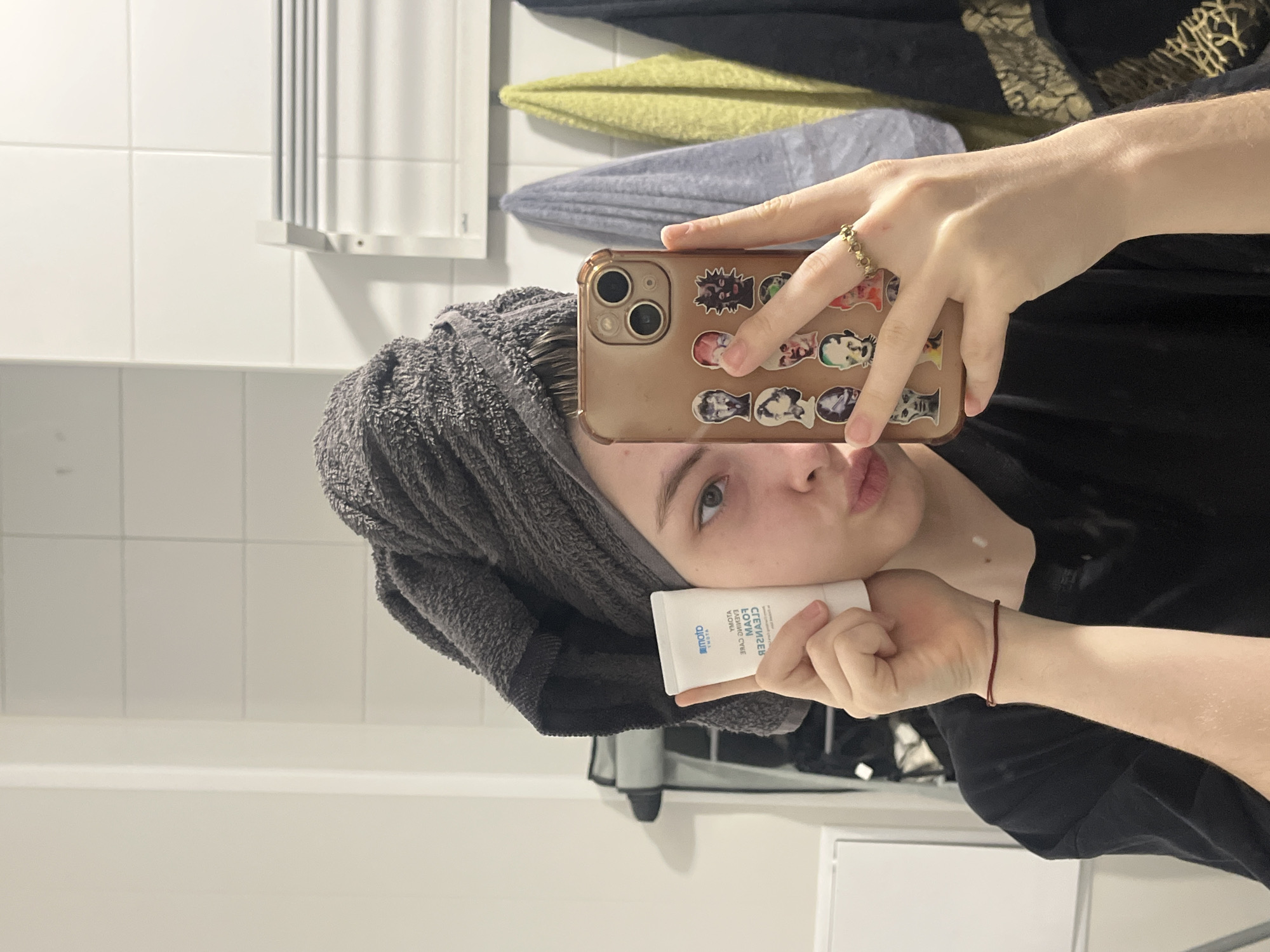 A person stands in a bathroom, taking a selfie in the mirror. The person has a grey towel wrapped around their head and holds a white tube of "YMOTA Foam Cleanser" in their left hand. They use a phone with a rose-gold case adorned with stickers to capture the image. The bathroom features white tiles, a towel rack, and several towels hanging on it. The person is wearing a black shirt. A ring is visible on their right hand, and a red bracelet is on their left wrist. The location in the city of Saint Petersburg, Russia, cannot be determined from this image.