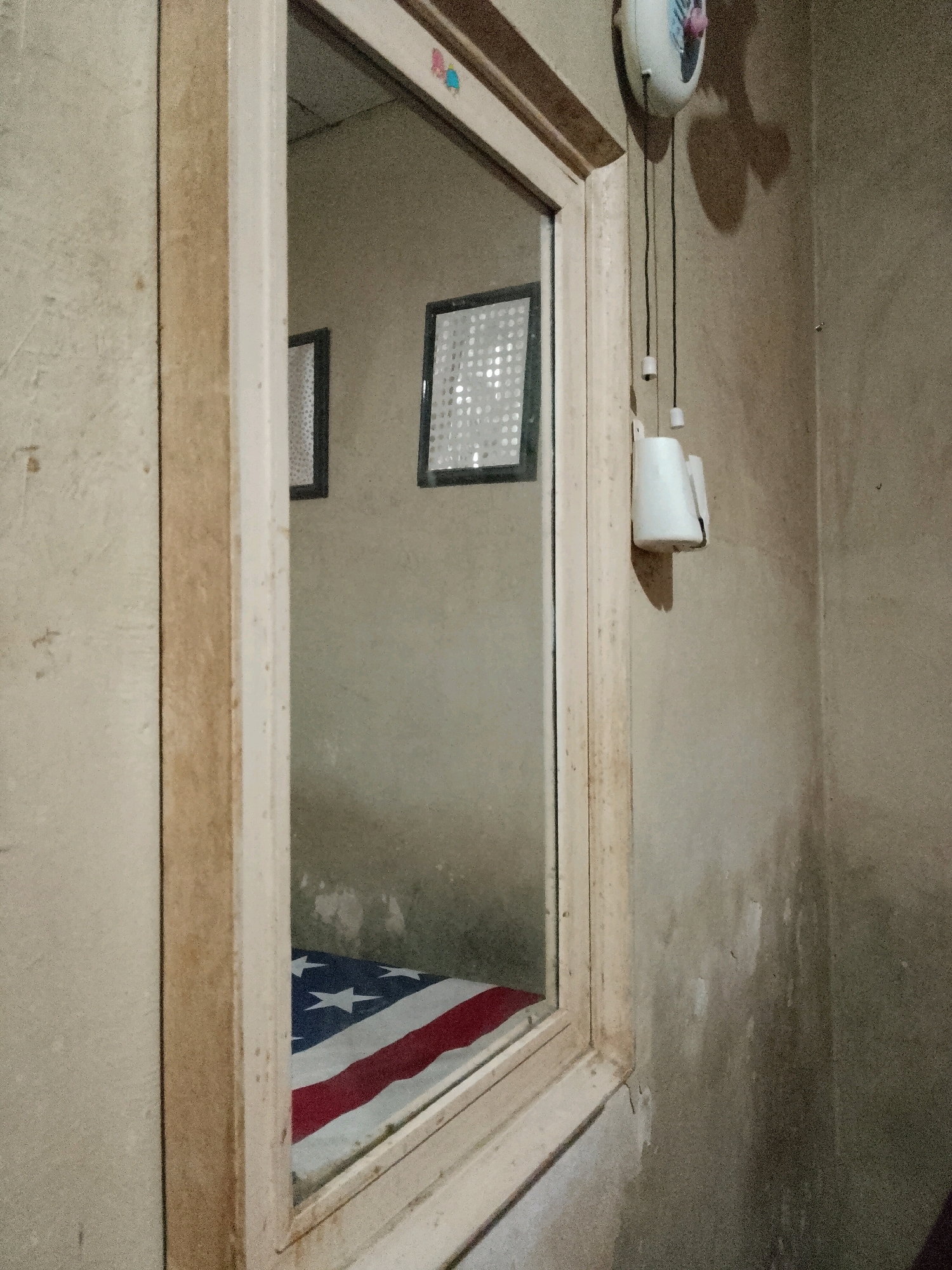 The image shows an interior scene, likely a bedroom, with a prominent wooden-framed mirror taking up the central portion of the frame. The mirror reflects an American flag draped over a surface, possibly a bed. To the right of the mirror, a white electrical device with pull cords hangs on a textured, neutral-colored wall. Above the mirror, a small clock or timer is visible. The overall impression is one of a sparsely decorated, possibly older, living space. The lighting is dim, creating shadows and a muted color palette. The image does not provide enough visual information to determine the exact location in Surabaya, Indonesia, or the presence of any people.