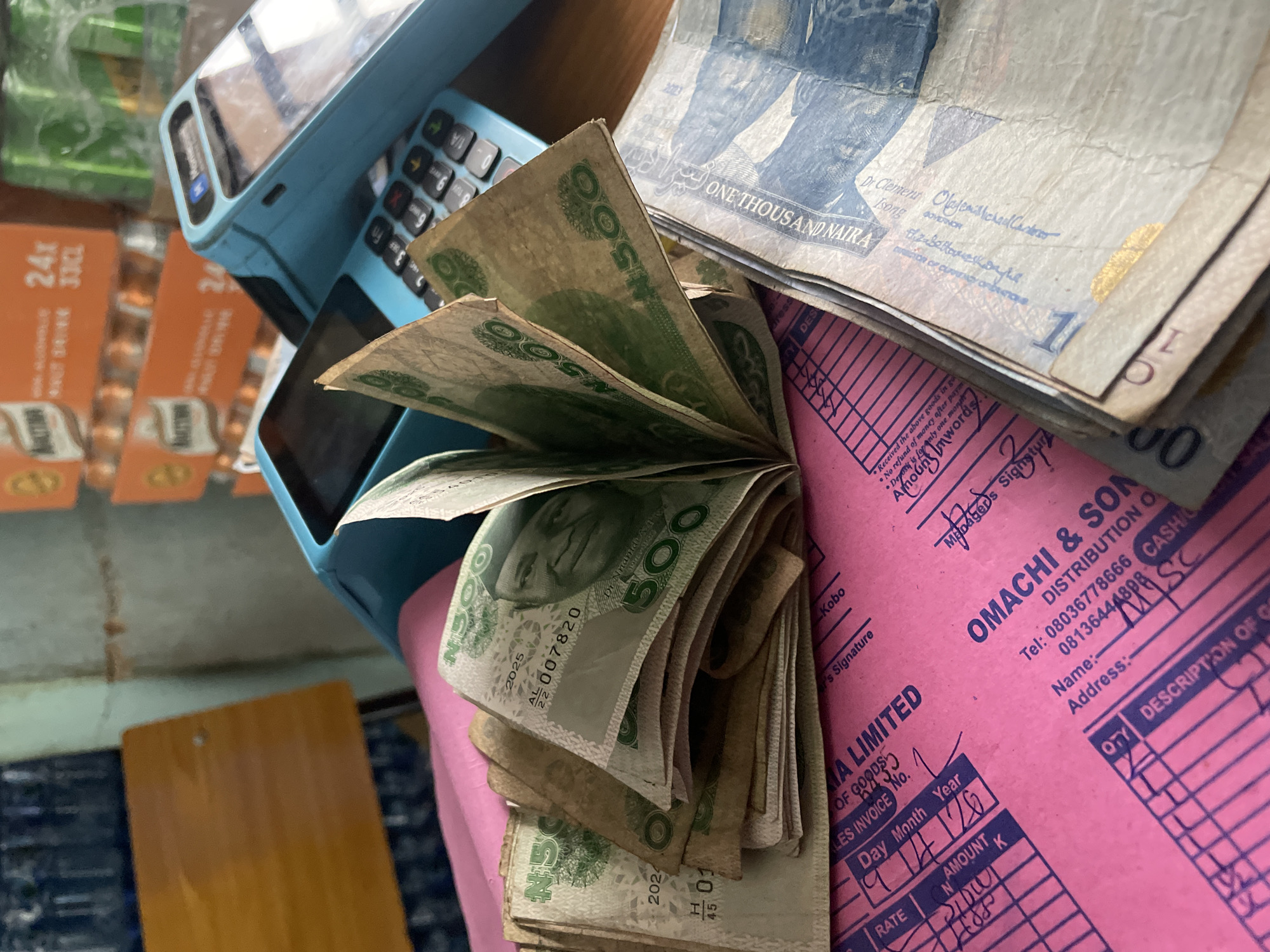The image shows a close-up view of Nigerian currency (Naira) and business-related items. There are stacks of ₦500 and ₦1,000 Naira notes. The ₦500 notes are partially fanned out, revealing the portrait of Dr. Nnamdi Azikiwe on some of them. The ₦1,000 notes are stacked, showing the image of Dr. Clement Isong. Adjacent to the money, there is a blue point-of-sale (POS) machine. The foreground includes a pink invoice or receipt booklet from "OMACHI & SONS DISTRIBUTION O..." with handwritten details on the top sheet. A partially visible box of Maltina drinks sits in the top left corner of the frame. The location is unknown, but given the local currency and invoice in the image, it can be said that this scene is most likely in Nigeria.