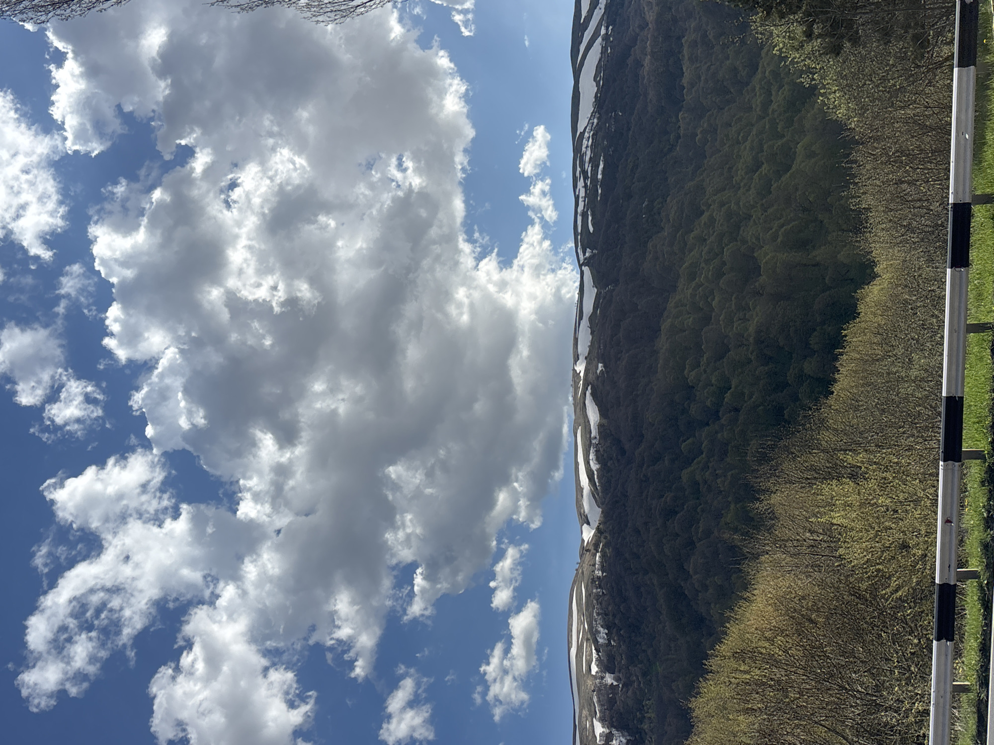 The image depicts a landscape, likely taken in a mountainous area. A large cloud formation occupies the left side of the frame, contrasting with a clear blue sky. On the right, a mountain covered in dense forest stretches upwards, with patches of snow visible near the peak. A safety rail is seen on the right side of the image. Given the query specifying Dilijan, Armenia, the image most likely depicts a mountain and landscape in that region.