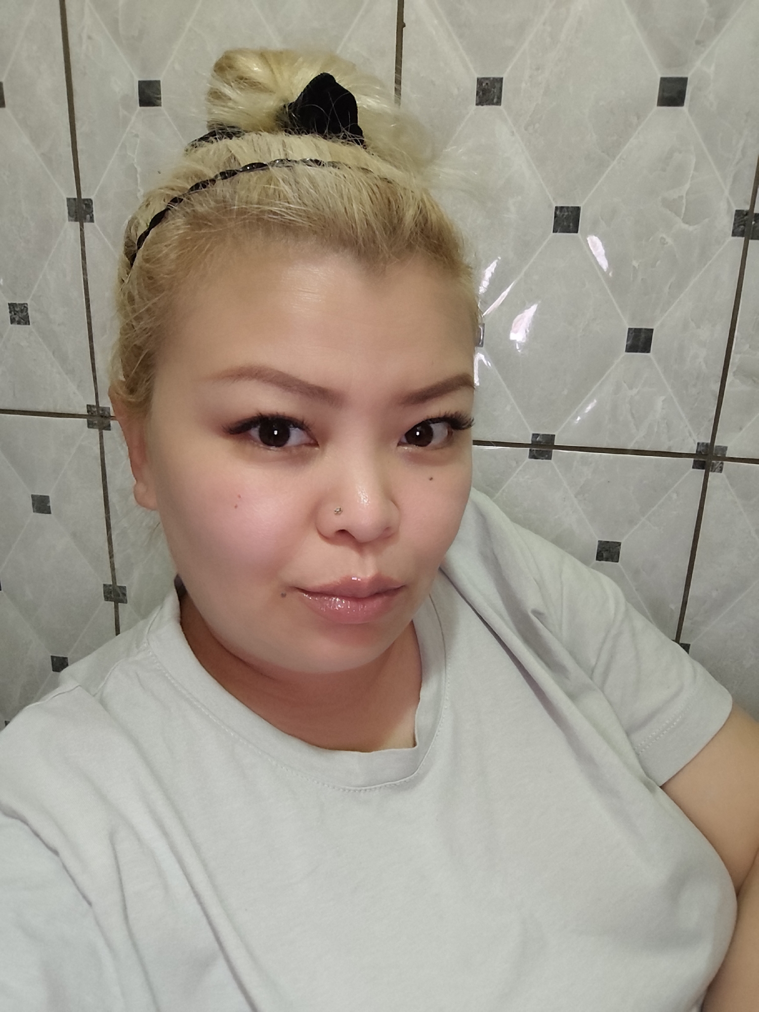 A close-up selfie shows a fair-skinned woman with blond hair pulled up in a bun, secured with a black headband. She is wearing a light-colored t-shirt. The background is a tiled wall with a geometric pattern of gray and black squares. The location is a room with tiled walls in the city of Unknown, Unknown.
