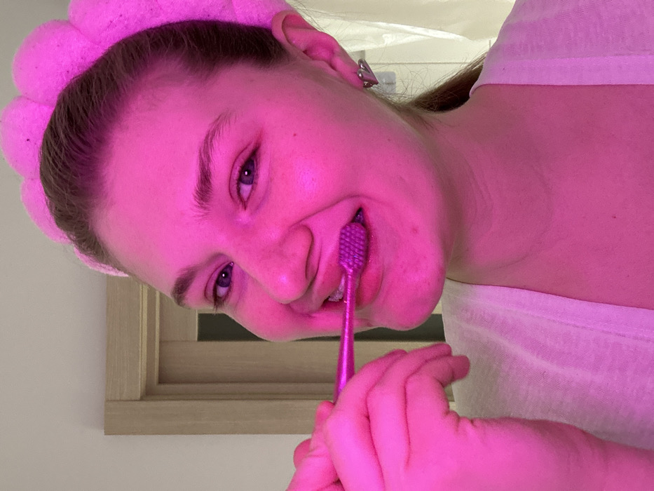 The image captures a close-up, rotated view of a young person, likely female, in a brightly lit, predominantly pink environment. The subject's head is tilted significantly, with the top of her head towards the left side of the frame.
The person has fair skin, which appears magenta due to the strong pink lighting. She has blue or violet-colored eyes and light-colored eyebrows. Her hair, which appears to be dark blonde or light brown, is pulled back, and she is wearing a fluffy, pink, flower-shaped headband that sits across her forehead and covers the top of her head. A silver or metallic earring with a geometric design is visible on her right ear (viewer's left).
She is in the process of brushing her teeth with a matching bright pink or magenta toothbrush. Her mouth is slightly open, revealing some teeth, and the bristles of the toothbrush are inside her mouth. Her right hand, also bathed in pink light, holds the toothbrush. She has a slight, closed-mouth smile, suggesting a neutral or pleasant expression.
The person is wearing a light-colored top, possibly white, which is visible on the right side of the frame. The background appears to be a simple light-colored wall. In the lower-left corner, part of a light-colored wooden frame, possibly from a window or mirror, is visible. The overall scene is dominated by the intense pink lighting, giving the entire image a vibrant, monochromatic feel. There is no visible text in the image.