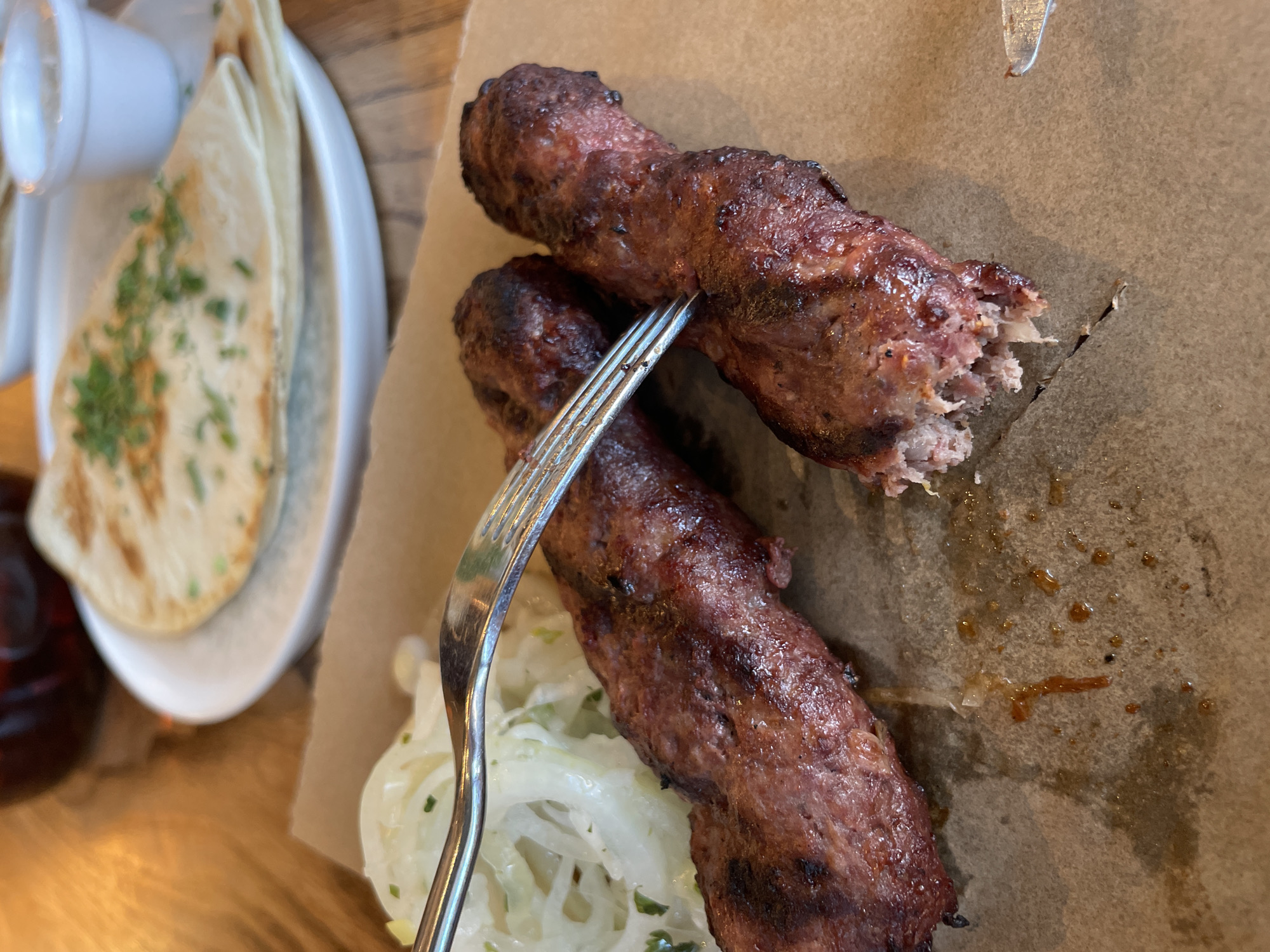 A close-up, overhead shot shows two grilled meat links, appearing to be sausages or kebabs, on a piece of brown parchment paper. One link is whole, while the other has been partially eaten, with its interior visible. A fork is positioned between the two links, with its tines partially inserted into the whole link. To the left of the meat, there is a serving of thinly sliced white onions, possibly dressed with a light sauce. In the background, out of focus, is a white plate with folded flatbread, sprinkled with chopped green herbs. A small white ramekin containing a white dipping sauce is also visible on the plate. A dark liquid, likely a beverage, is in a glass or cup at the top of the frame. The setting appears to be a restaurant table, with a wooden surface visible. The lighting is natural and warm.