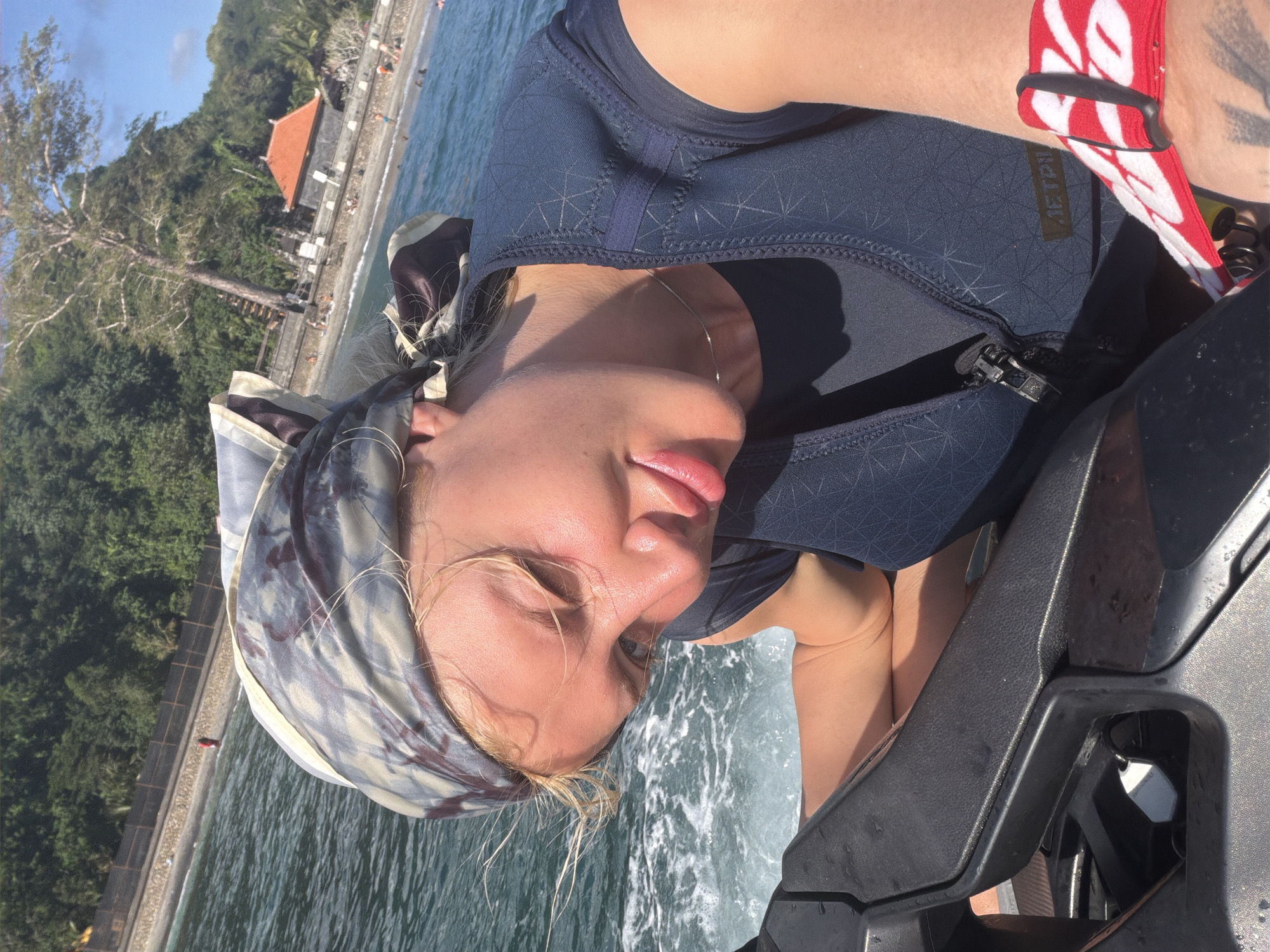 A close-up, selfie-style shot features a blonde woman with light skin wearing a patterned headscarf and a dark blue vest. She is positioned on a jet ski, with the water and a tropical coastline visible in the background. The woman is looking down and to her left, with her eyes partially closed. Her lips are slightly parted and appear to have lipstick on. A red and white patterned band is visible on her left wrist. The background shows a beach with several people, lush green trees, and a traditional Balinese structure with an orange roof. The water is choppy and appears to be moving. This scene is likely located at a beach or coastal area in Nusa Penida, Indonesia, given the distinct architectural style of the building and the tropical vegetation.