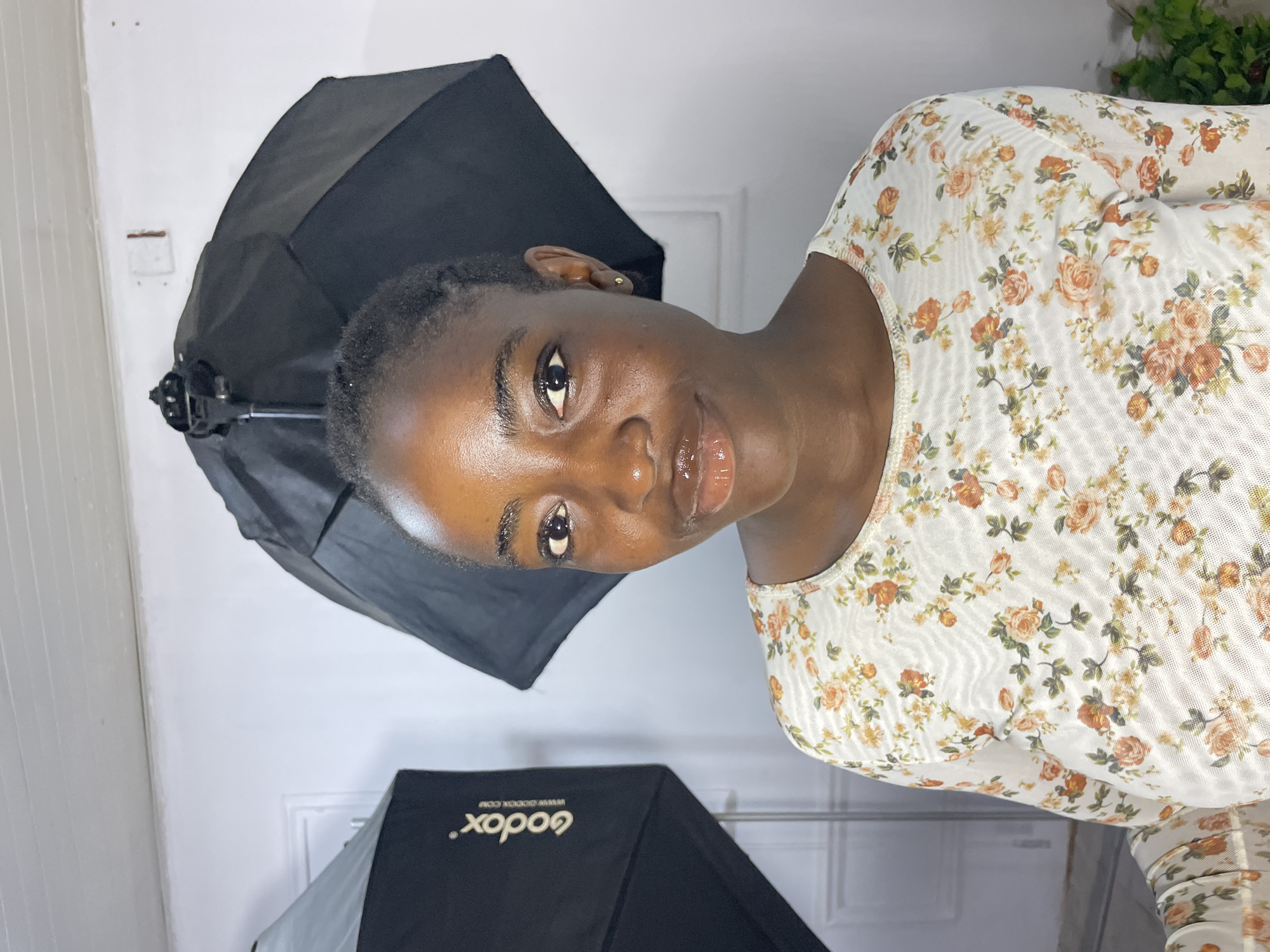 A young Black woman is photographed from the chest up, looking directly at the camera. She has dark skin, brown eyes, and her hair is pulled back. She is wearing a white, long-sleeved top with a floral print featuring orange and peach roses and green leaves. She has on lip gloss and a small stud earring in her left ear.
In the background, to the left, is a black softbox with the "Godox" logo visible. To the right and behind her is a black umbrella-shaped photographic reflector or softbox. The background wall is white and appears to be indoors. The lighting suggests a studio setting. The image quality is clear, and the subject is well-lit. There is no indication of the specific location within Jos, Nigeria.