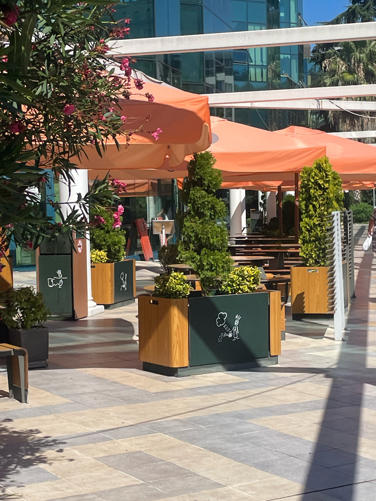 The image depicts an outdoor seating area with orange umbrellas providing shade. In the foreground, there are several large planters containing green shrubs and trees. One planter features a dark green panel with a white cartoon illustration of a person watering plants with a watering can, with steam or mist rising from the plants. Another planter has a similar illustration depicting a figure with a broom and dustpan. The ground is paved with large, light-colored square tiles arranged in a pattern. In the background, there are tables and chairs, suggesting a cafe or restaurant. A modern building with glass windows and a light-colored facade is visible behind the seating area. Lush green trees and palm trees are also present in the background. The lighting suggests a sunny day, with shadows cast on the ground. The overall scene conveys a clean and pleasant outdoor urban environment.