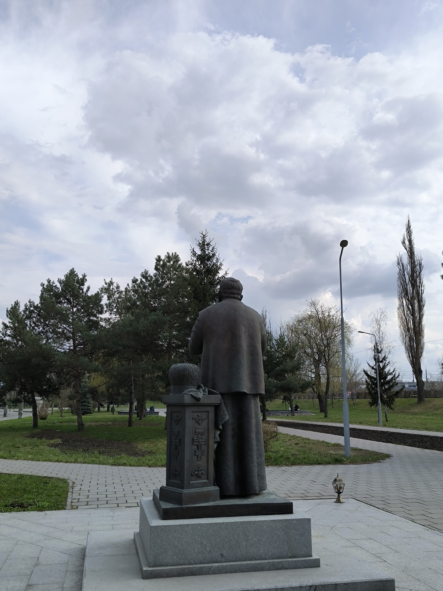 The image shows a bronze statue in a park in Vinnytsia, Ukraine. The statue depicts a man in a long robe with his back to the viewer. Next to him is a pedestal with a decorative crown on top. The statue is mounted on a multi-tiered stone base. Behind the statue are various trees and a pathway. A streetlight stands to the right of the statue. The sky is overcast with numerous clouds.