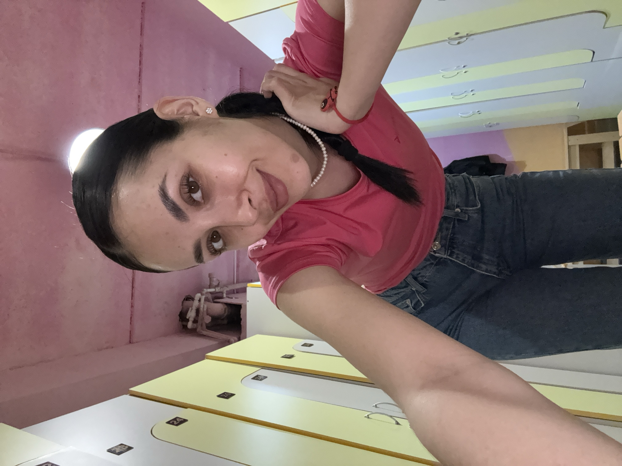 A woman with dark hair in a ponytail, wearing a pink shirt and jeans, is in a room with lockers. The room has pink and yellow walls and lockers with numbers on them. There is a medical device visible in the background. The location is Votkinsk, Russia.