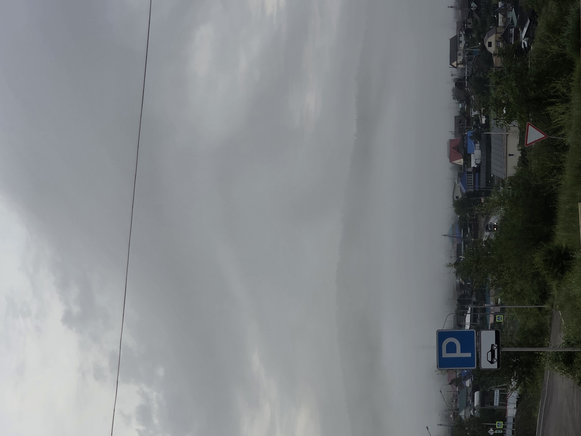 The image depicts a street scene in a village, likely in Chita, Russia, under overcast skies and fog. The dominant subjects are the residential houses with varying roof colors, interspersed with green trees and vegetation. A road is visible in the foreground, suggesting a transportation route.
Several traffic signs are present. A blue square sign with a white "P" indicates parking. Next to it, a rectangular sign shows a white silhouette of a car on a black background, possibly signifying a parking area for cars or a specific parking regulation. A triangular red and white sign, commonly used to indicate a "Give Way" or "Yield" warning, is also visible. Further down the road, smaller yellow signs with a triangle and an exclamation mark are placed, likely indicating further warnings or information for drivers. One of these signs has Cyrillic text that appears to read "СУХОДОЛ".
The atmosphere is hazy and diffused due to the fog, obscuring the distant view of the village and creating a somewhat muted color palette. The time of day is not clearly indicated but the gray, cloudy sky suggests it could be daytime. The overall impression is of a quiet, perhaps damp, day in a rural or semi-rural setting. There are no people clearly visible in the frame. The presence of parked vehicles in the background suggests this is an active area despite the weather.
