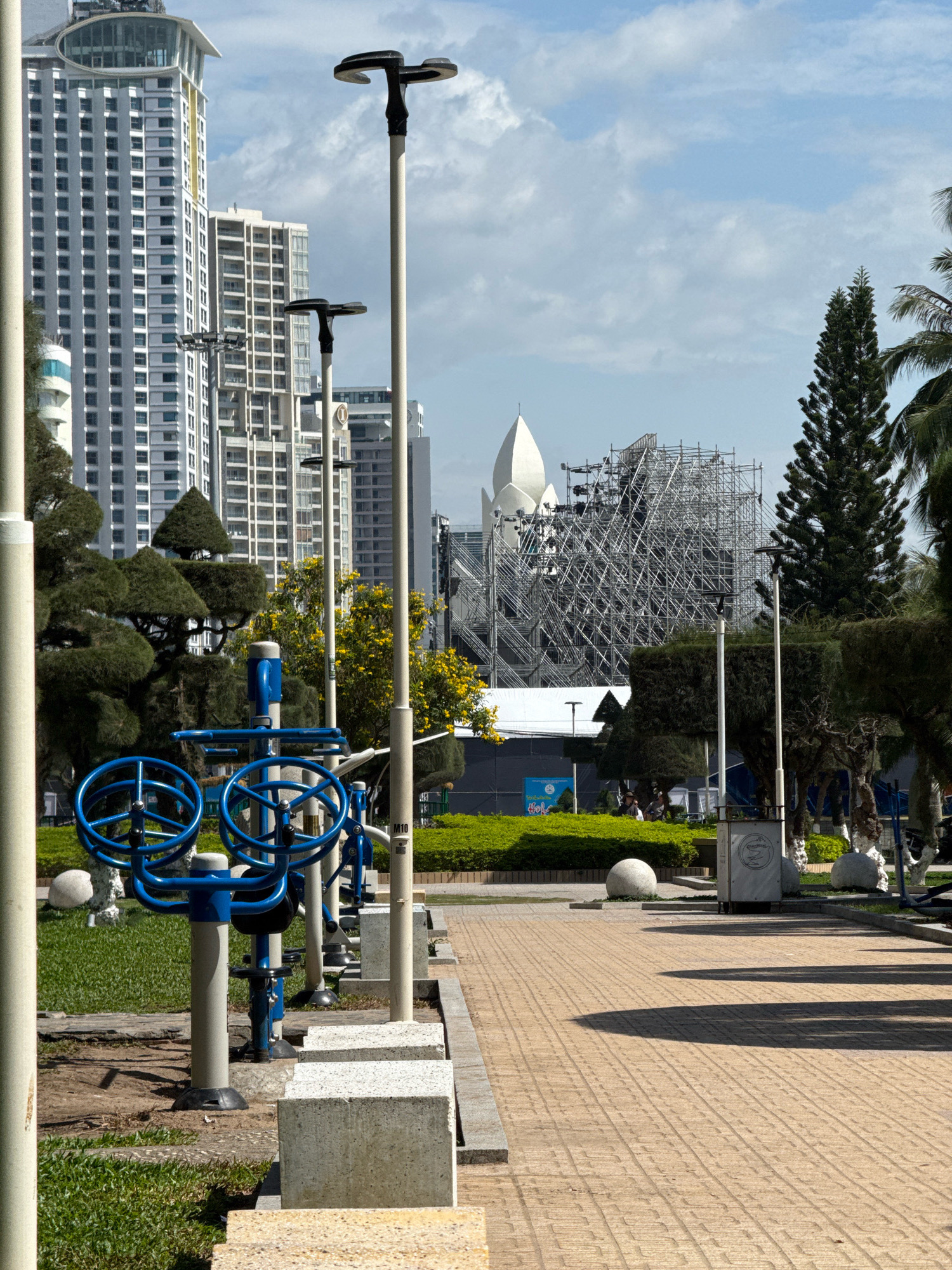 The image depicts an outdoor exercise area in Nha Trang, Vietnam, featuring blue exercise equipment with large circular handles positioned on a grassy area. A paved walkway with a textured surface runs alongside the grass, casting long shadows from the surrounding elements. In the background, several tall, modern buildings with a white facade and numerous windows dominate the skyline. Further back, a distinctive white, lotus-shaped building is visible, partially obscured by extensive scaffolding, indicating ongoing construction or renovation. Several lampposts are spaced along the walkway, and manicured trees and bushes contribute to the urban landscape. The sky is partly cloudy, with a clear blue expanse overhead.