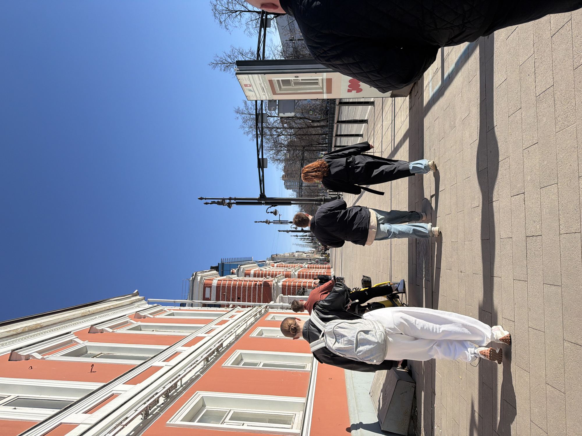 On a sunny day in Perm, Russia, a street scene unfolds with several people walking along a wide pedestrian walkway. The image is shot at an oblique angle, with a clear blue sky dominating the upper portion.
A series of buildings with red and white facades line one side of the walkway. The architecture is characterized by multiple windows and decorative molding. Street lamps are visible along the walkway.
Several people are walking in the same direction along the walkway. One woman has red hair and is wearing a black coat. A man in a dark jacket and jeans is walking behind her. Another person is with a bicycle, near a pedestrian wearing white clothing. A person wearing a black jacket is partially visible in the foreground.
The walkway itself is paved with rectangular stones. Shadows cast by the people and objects indicate the direction of sunlight.