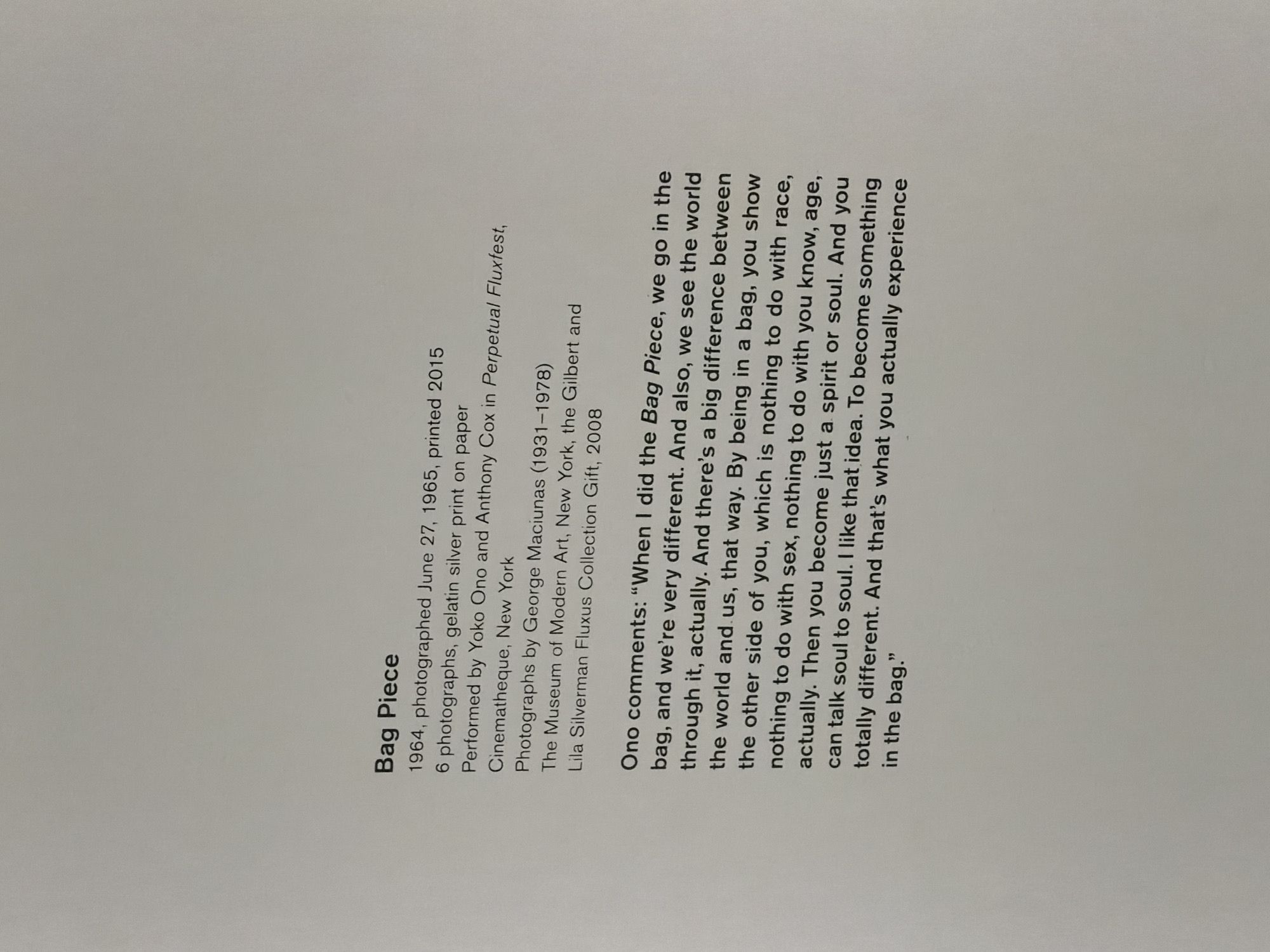 This image shows the text associated with an artwork called "Bag Piece". The text provides information about the piece, including that it was photographed in 1964, printed in 2015, and consists of 6 photographs, gelatin silver print on paper. It was performed by Yoko Ono and Anthony Cox in Perpetual Fluxfest, Cinematheque, New York. The photographs were taken by George Maciunas. The piece belongs to the Museum of Modern Art, New York, and is part of the Gilbert and Lila Silverman Fluxus Collection Gift, 2008. The text also includes a quote from Yoko Ono about the piece.