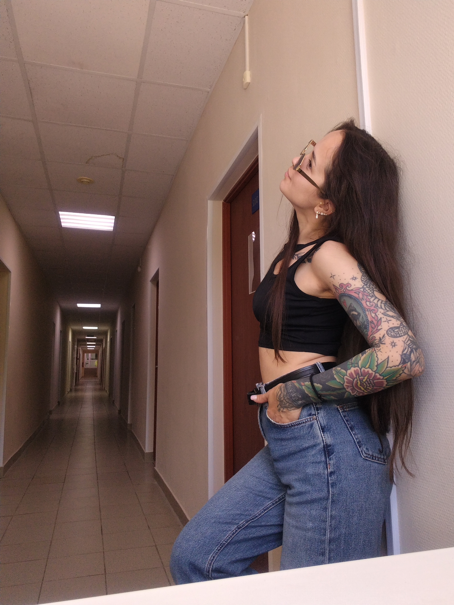 A woman with long dark hair, wearing gold-rimmed glasses, a black crop top, and blue jeans, is posing in a long indoor hallway in Chaikovsky, Russia. She leans casually against a light-colored wall, looking upwards and slightly to her right. Her right hand is tucked into her jeans pocket. Both of her arms are extensively covered in colorful full-sleeve tattoos featuring intricate floral and traditional designs in shades of red, green, and blue. She also wears multiple earrings in her left ear.
The hallway features a suspended ceiling with square, textured tiles, one of which appears slightly sagged or damaged near the foreground. Rectangular fluorescent light fixtures are spaced along the ceiling, illuminating the corridor and creating a sense of depth as it recedes into the distance. The floor is covered with light-colored square tiles. Numerous doors, some dark brown and others lighter, line both sides of the hallway, suggesting access to various rooms or offices. The overall scene is brightly lit by artificial light, and the mood is calm. Very faint, indistinct blue text, possibly "SYO," is barely visible on a brown door to the woman's left.