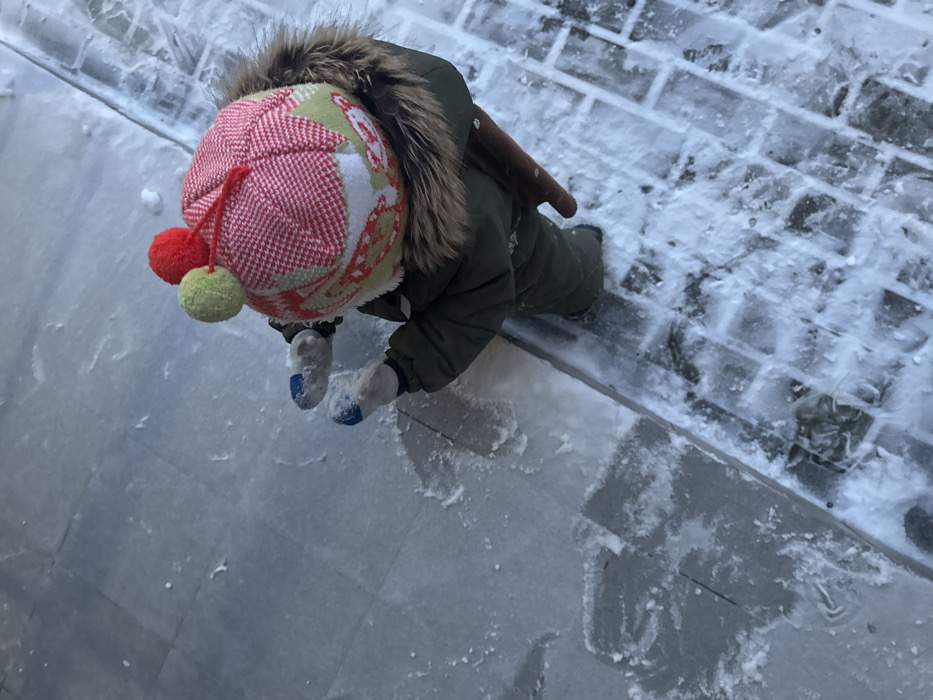 An overhead photograph captures a young child dressed in warm winter attire, crawling on a snow and ice-covered ground in Odintsovo, Russia. The child wears an olive green winter jumpsuit with a fur-lined hood and a brightly patterned hat featuring red, light green, and white sections, topped with two prominent pom-poms in red and light green. Their hands are covered in light-colored mittens with blue tips. A brown object, possibly part of a sled or a toy, is visible resting on the child's back.
The ground surface is largely covered in a thin layer of snow and ice, showing textures indicative of cold winter weather. The paving beneath is split into two distinct areas: a smoother, lighter grey section in the foreground, and a darker, textured brick-like pattern further back, both partially obscured by the frozen precipitation. The scene suggests a daytime setting, likely under overcast skies, with diffused light. The child is actively exploring or playing on their hands and knees in the snowy environment. No visible text is present in the image.