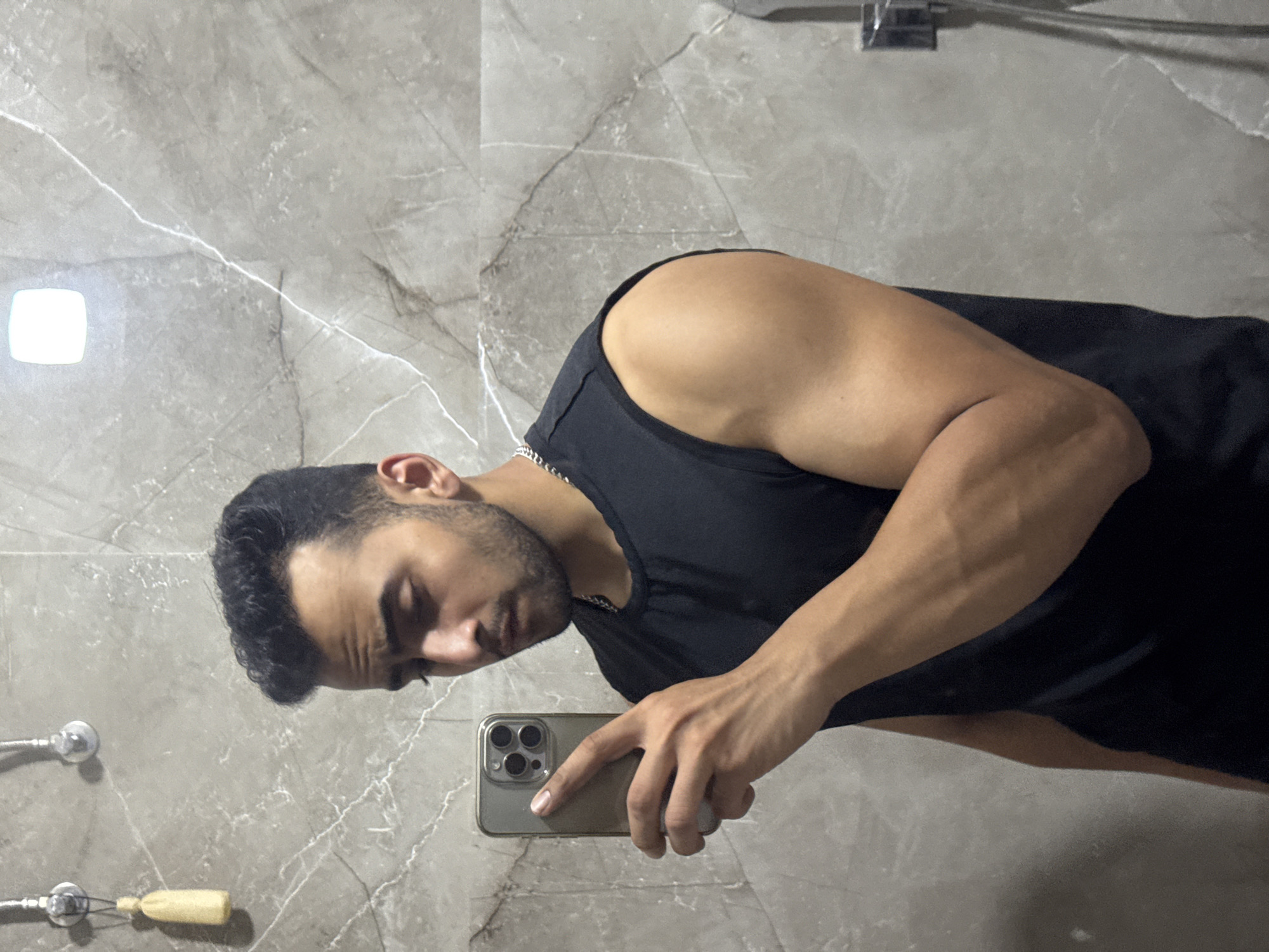 A man stands in a bathroom with gray marble walls, taking a selfie with a gold iPhone. He is wearing a black tank top and a silver chain. A light fixture is visible on the wall, along with two towel racks. The location is not specified, and Kolkata, India, is not mentioned or indicated.