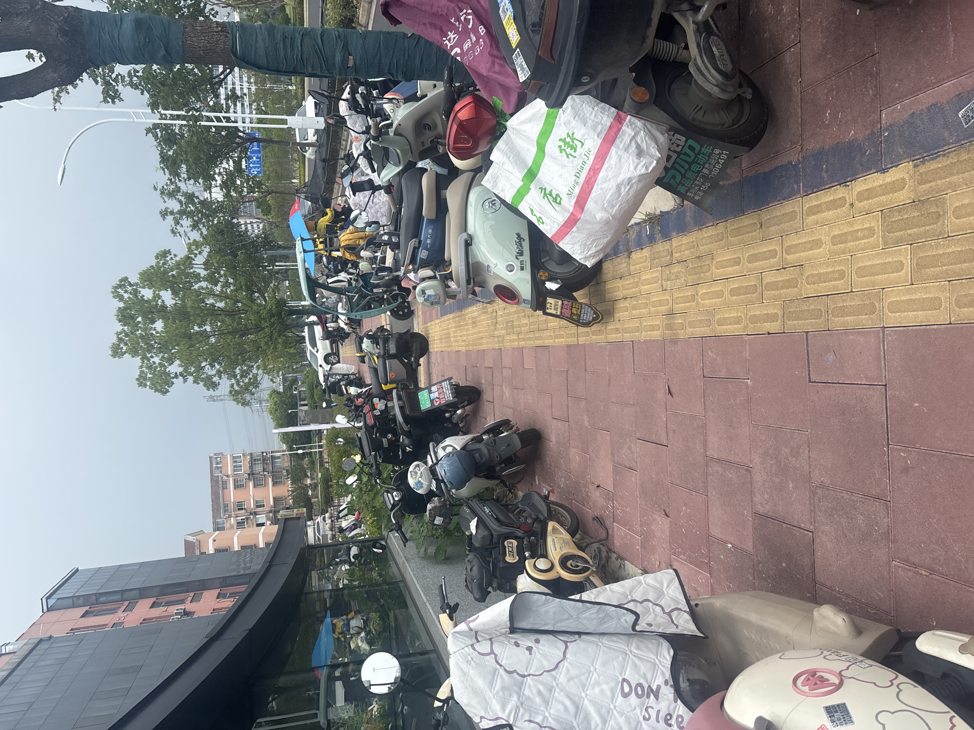 In Suzhou Industrial Park, China, a sidewalk is lined with numerous parked electric scooters and motorcycles. The scooters are mostly white, grey, and blue, with some covered by protective cloths. A few individuals are visible, one wearing a yellow jacket and blue umbrella, suggesting rainy weather. The sidewalk is paved with red and yellow tiles. In the background, a modern building with dark grey and pink facades is present, along with trees and a street lamp. The scene appears to be an outdoor parking area for personal transportation.