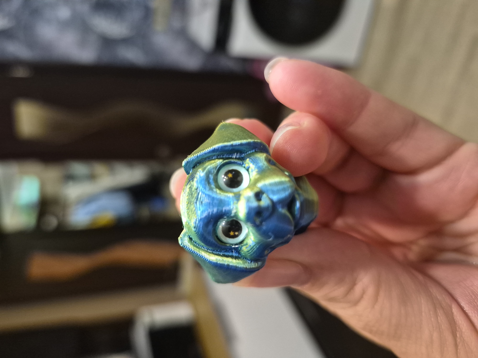 A close-up, slightly blurry image shows a person's hand holding a small, multi-colored 3D-printed figurine. The figurine appears to be a stylized creature with two large, expressive eyes, each with a black pupil and a yellowish, star-shaped reflection. The creature has a hat-like structure on its head, colored in shades of green and blue, with a textured brim. The body of the figurine is also a blend of blues and greens, with visible layers from the 3D printing process. The hand holding the figurine is a pale skin tone, with the fingers gently cupping the object. The background is out of focus, but suggests an indoor setting with some shelves or furniture visible. No text is present in the image. The lighting appears to be artificial, creating soft shadows. The overall impression is of a whimsical, handcrafted item being presented or examined.