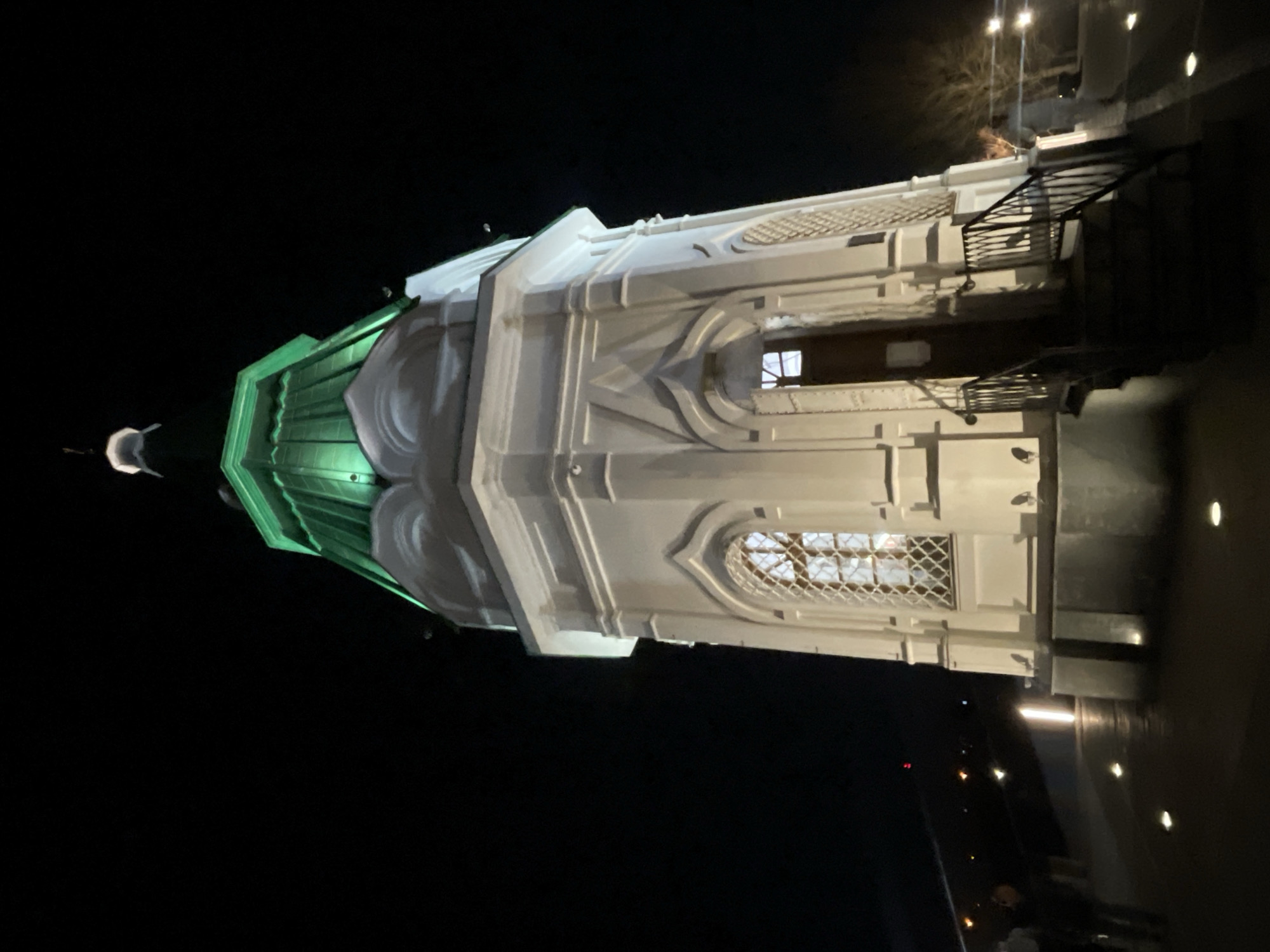 The image depicts the Paraskeva Pyatnitsa Chapel in Krasnoyarsk, Russia at night. The chapel has a green dome with a spire at the top and white walls with intricate architectural details and windows. There is also a metal railing at the entrance of the chapel. The scene is dark with some illumination highlighting the chapel and the surrounding area, which shows a pathway and people standing near the edge of the pathway.