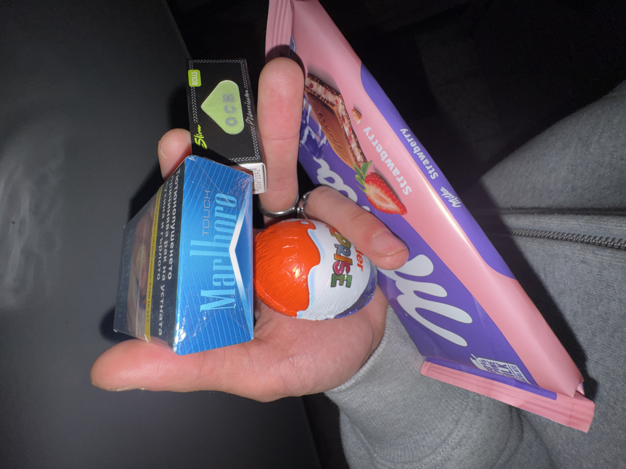 A human hand holds a collection of items. From left to right: A blue package of Marlboro Touch cigarettes with Bulgarian text. Above the cigarettes is a green and black package of OCB Slim Rolls rolling papers. In the center is an orange and white Kinder Surprise egg. To the right is a pink and purple Milka Strawberry chocolate bar. The hand appears to be resting against a grey sweatshirt. The image is taken indoors, possibly in a dark room or at night. There are no specific indicators of Sofia, Bulgaria.