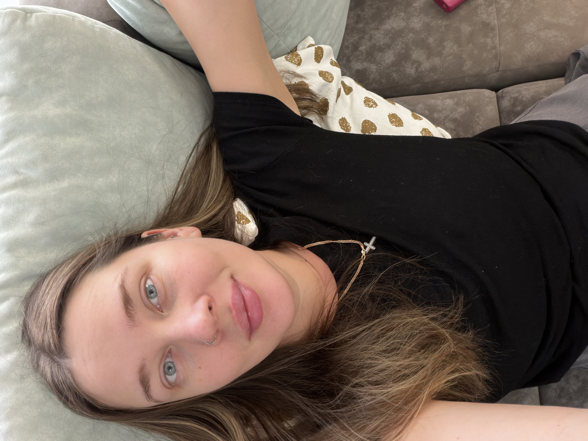 A woman with light skin, blue eyes, and brown hair with blonde highlights is lying down. She is wearing a black top and a gold necklace with a cross pendant. She has a nose ring in her left nostril. Her head rests on a light green pillow. Behind her is another pillow with gold spots on a white background. She appears to be lying on a gray couch. The location is not discernible from the image.
