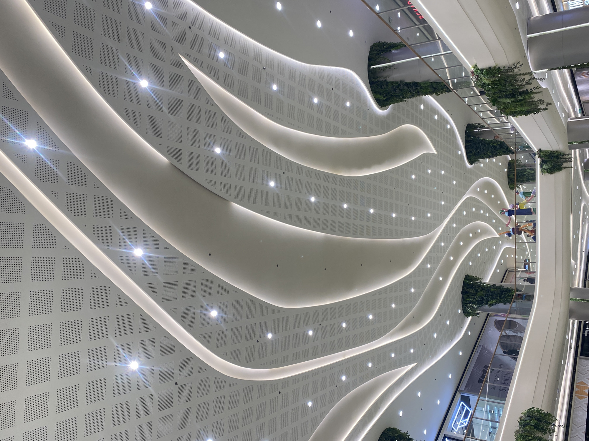 The image shows the interior of a modern shopping mall with a distinctive curved ceiling design. The ceiling is white and features a grid of square perforations, with embedded circular lights. Dramatic, illuminated white curves sweep across the ceiling, creating a sense of organic flow. Tall cylindrical pillars are visible, wrapped in hanging greenery, adding a natural element to the architectural design.
The mall appears to be multi-leveled, with balconies and railings visible in the background. People are sparsely distributed throughout the visible areas, suggesting a quiet or off-peak time. On one of the upper levels, a display with mannequins or figures dressed in colorful clothing is present, seemingly part of a promotion or decoration.
In the background, signage for "SUNGLASS HAVEN" and "D ENIN" is visible, indicating retail establishments within the mall. The overall aesthetic is contemporary and clean, with a focus on light and flowing forms.
Based on the architectural style and branding visible, this is likely SM Lanang Premier, a major shopping mall located in Davao City, Philippines. SM Lanang Premier is known for its distinctive contemporary architecture and its integration of natural elements like the hanging greenery.