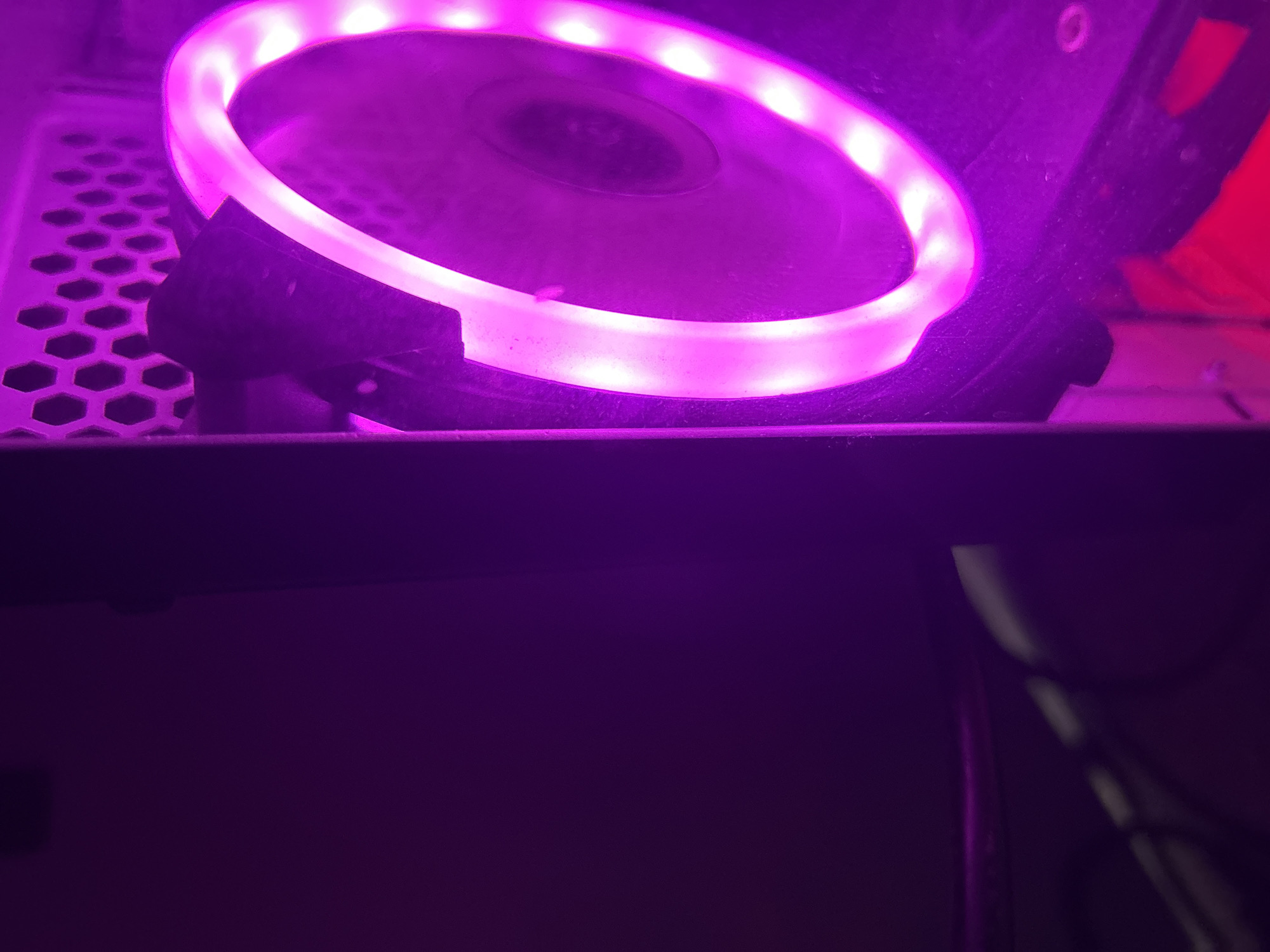A close-up shot of a circular computer fan with a ring of pink LED lights is situated inside a computer case. The fan is surrounded by black plastic housing. A metal honeycomb grill is partially visible in the background. The lower part of the image shows the dark interior of the computer case, with a cable running along the right side. The scene is dimly lit, emphasizing the bright glow of the LED lights. The location is unspecified.