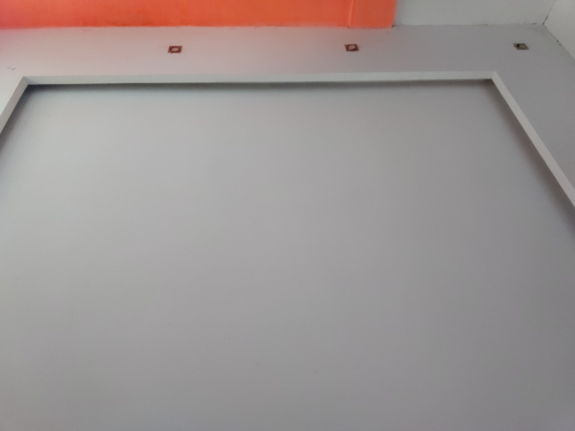 This image captures an upward-facing interior view of a room, primarily showcasing a wall and ceiling. The central focus is a large, light gray or off-white wall, which features a prominent white rectangular molding or frame. This frame creates a border effect, suggesting a defined space on the wall, possibly for artwork or a television.
Above the main wall, the ceiling is predominantly white, but a distinct band of vibrant orange or terra cotta color runs horizontally along the junction where the wall meets the ceiling. Several small, square-shaped recessed fixtures, appearing dark and empty, are visible in the white ceiling area directly above the orange band and further into the white ceiling. These fixtures are symmetrically placed.
The lighting is even, suggesting either diffused natural light or well-distributed artificial light within the indoor setting. There are no people, activities, or visible text in the frame. The image offers a clean, architectural perspective of a modern interior, highlighting the contrasting colors and structural details of the wall and ceiling.