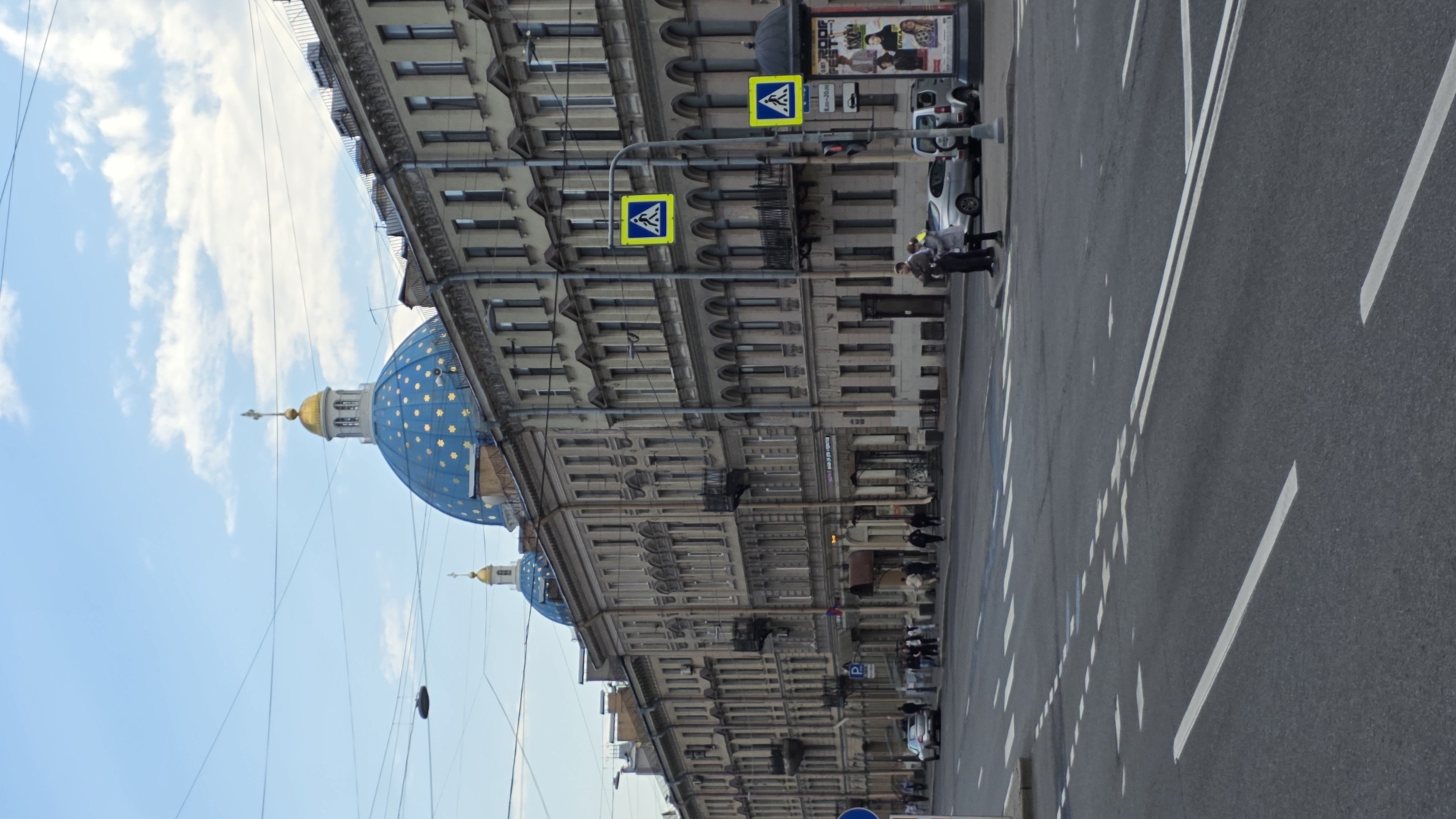 This image depicts a street scene in Saint Petersburg, Russia, featuring the Trinity Cathedral. The blue dome with gold stars of the cathedral is visible on the left side of the image. Adjacent to the cathedral is a multi-story building with rows of windows and ornate architectural details. Two yellow pedestrian crossing signs are positioned on poles near the building.
On the right side is a paved street with white lane markings. Several cars are parked along the street, and a few pedestrians are walking or standing near the parked vehicles. The sky is partially visible, showing light blue with some clouds.