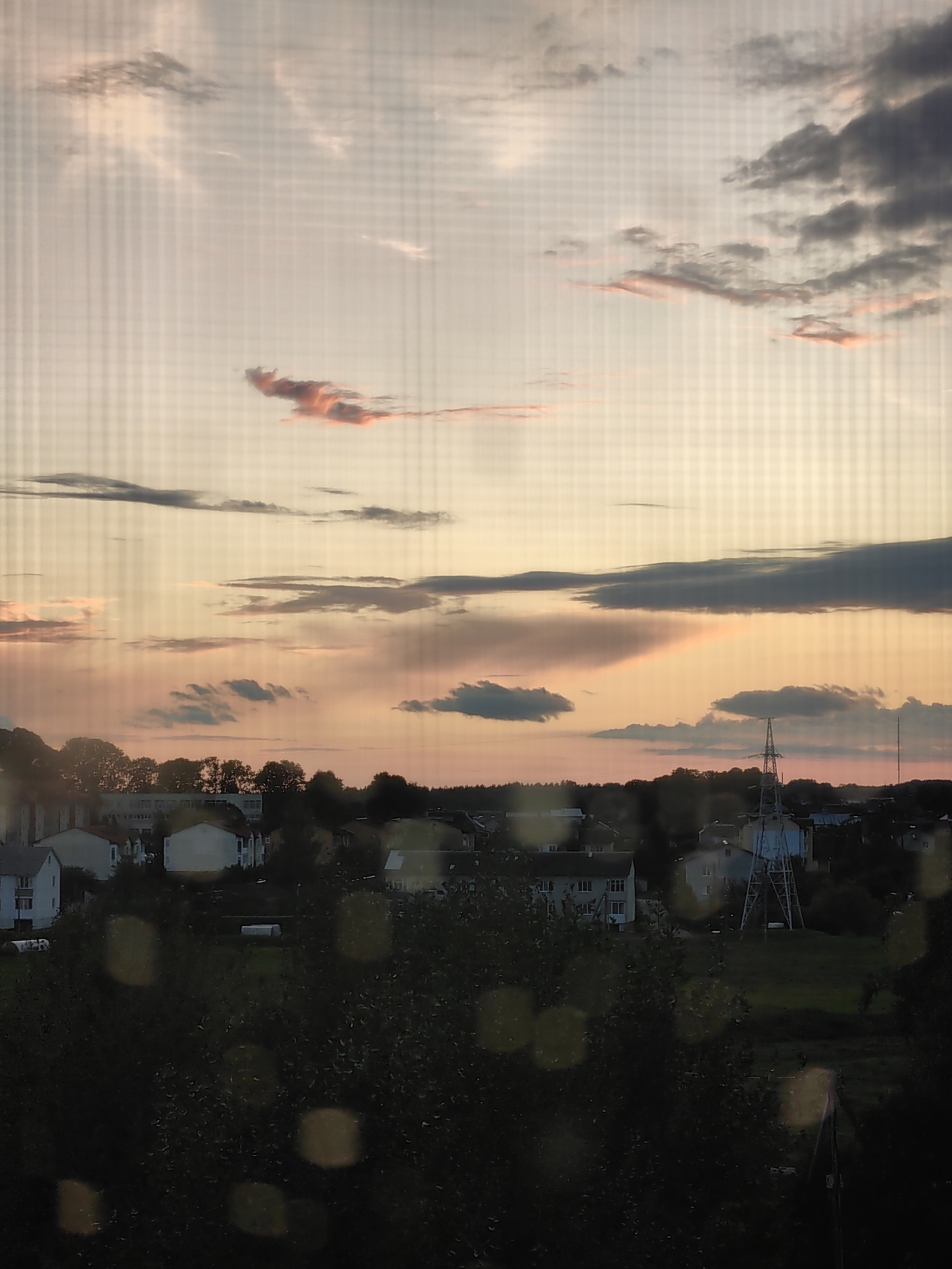 The image captures a sunset over a residential area in Myadzyel, Belarus. The sky is filled with soft clouds tinged with pink and orange hues. Below the sky, a collection of houses with white and beige facades are visible, interspersed with trees and greenery. A prominent power line tower stands in the mid-ground, suggesting the presence of electricity infrastructure. The foreground is dominated by dark foliage, possibly trees, with some out-of-focus circular light flares. The overall scene depicts a peaceful, twilight ambiance in a suburban setting.