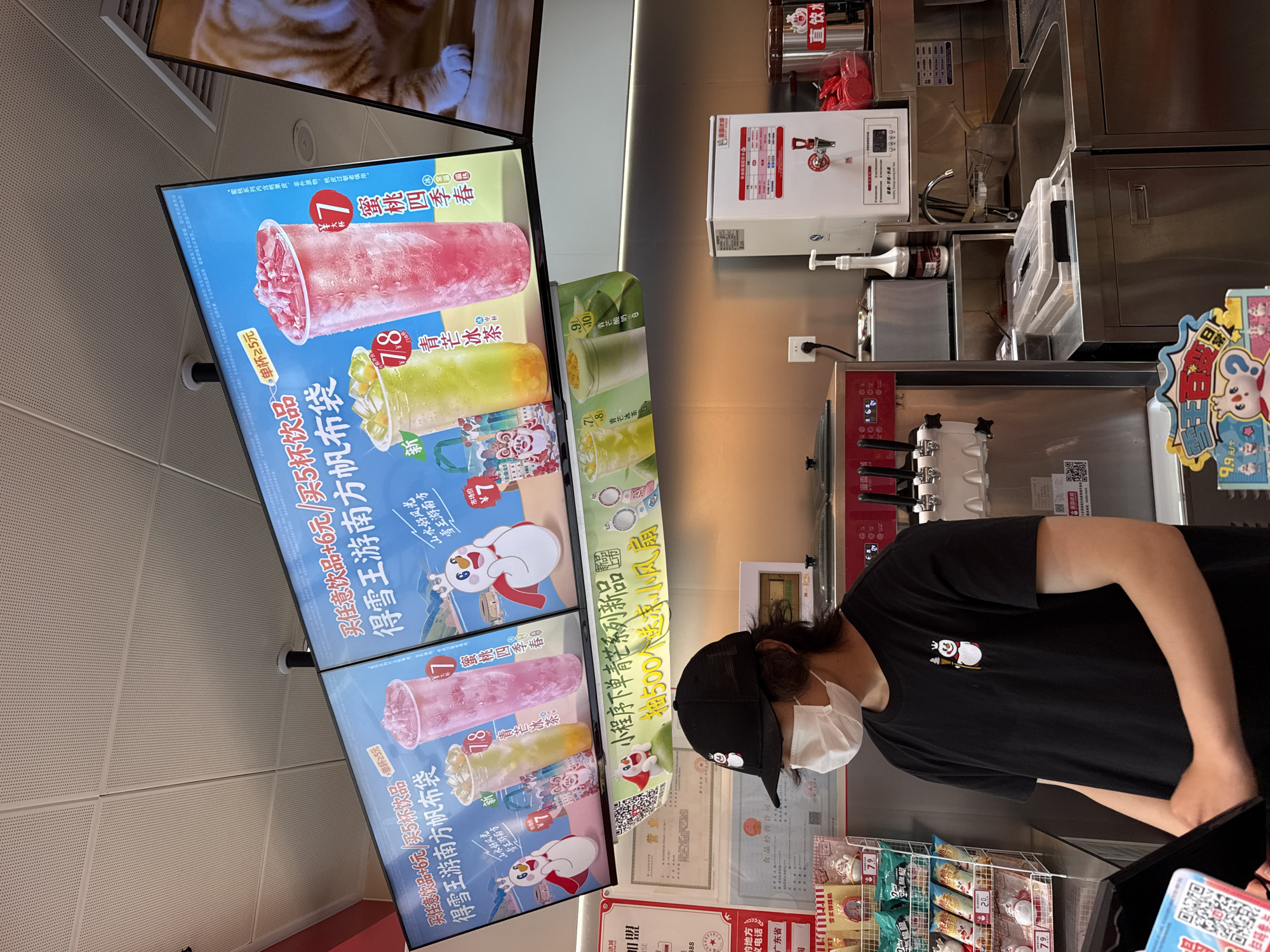 The image shows an indoor scene at a beverage store, likely in Huizhou, China. A person wearing a black t-shirt with a cartoon snowman logo and a black baseball cap, along with a white face mask, stands behind a counter, interacting with a point-of-sale system. The background features large digital menu boards displaying colorful advertisements for drinks, featuring cartoon characters and prices in Chinese Yuan. To the right, a large screen displays an image of a cat. Various beverage preparation equipment, including ice cream machines, dispensers, and sinks, are visible on the counter and in the background. Shelves stocked with packaged goods are also present. The overall environment appears to be a clean and modern retail space.