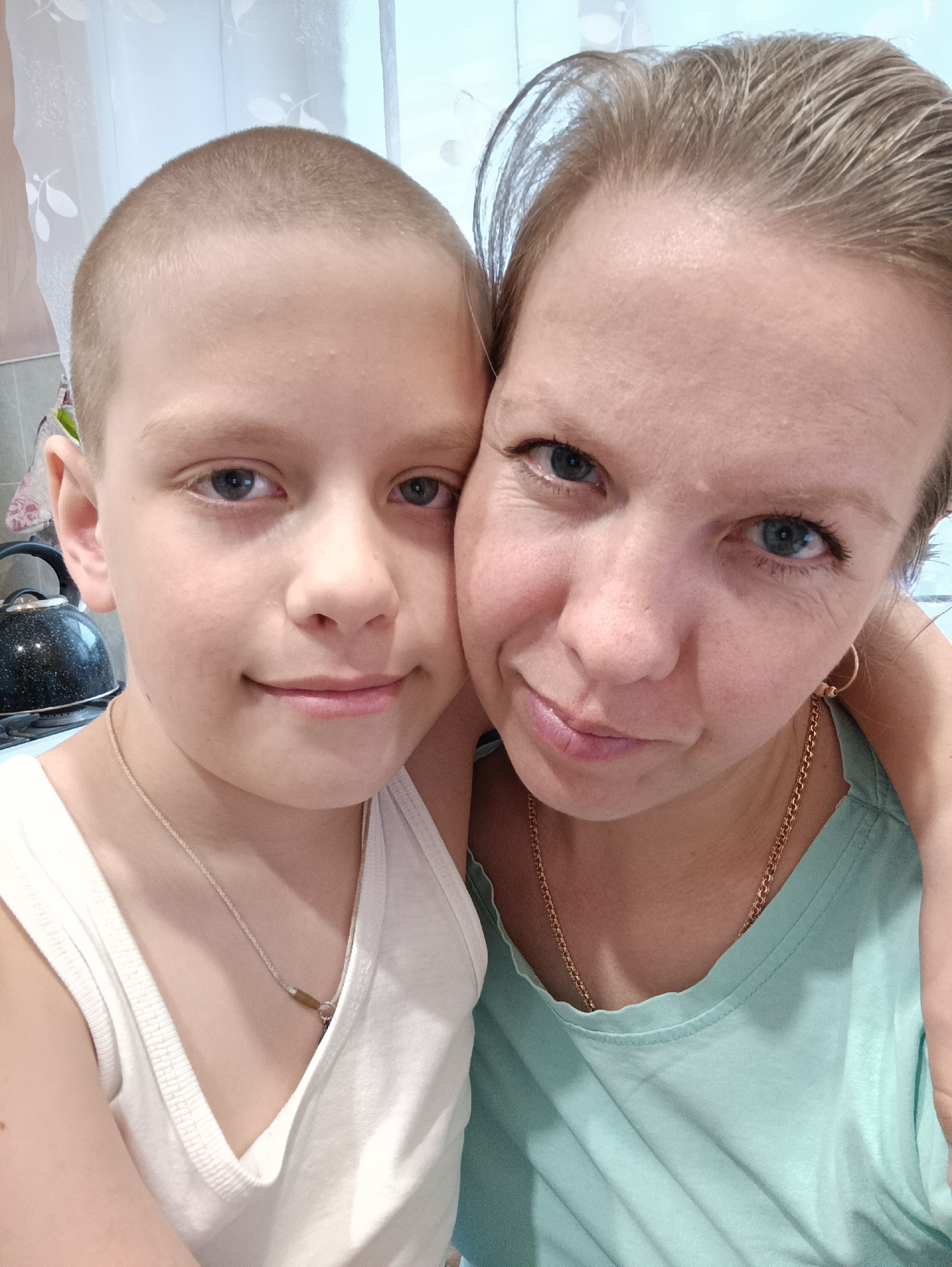 This close-up, informal portrait features an adult woman and a child, likely a boy, possibly a mother and son, posing together indoors in Chaikovsky, Russia. Both individuals are looking directly at the camera with gentle, serene expressions, conveying a sense of warmth and contentment.
The woman, positioned on the right, has light-colored hair pulled back and striking blue eyes. She is wearing a light green or teal top, a gold chain necklace, and a small gold hoop earring on her visible ear. Her arm is around the child, suggesting an embrace.
The child, on the left, has short, buzzed blonde hair and blue eyes. He is dressed in a white tank top and a light-colored cord necklace featuring a small charm.
The background elements indicate a domestic setting, likely a kitchen. To the left of the child, a black kettle rests on what appears to be a stovetop. Behind the subjects, a light-colored curtain with a subtle floral or leaf pattern is visible, suggesting natural light entering from a window. The soft lighting indicates it is daytime. There is no visible text in the image.