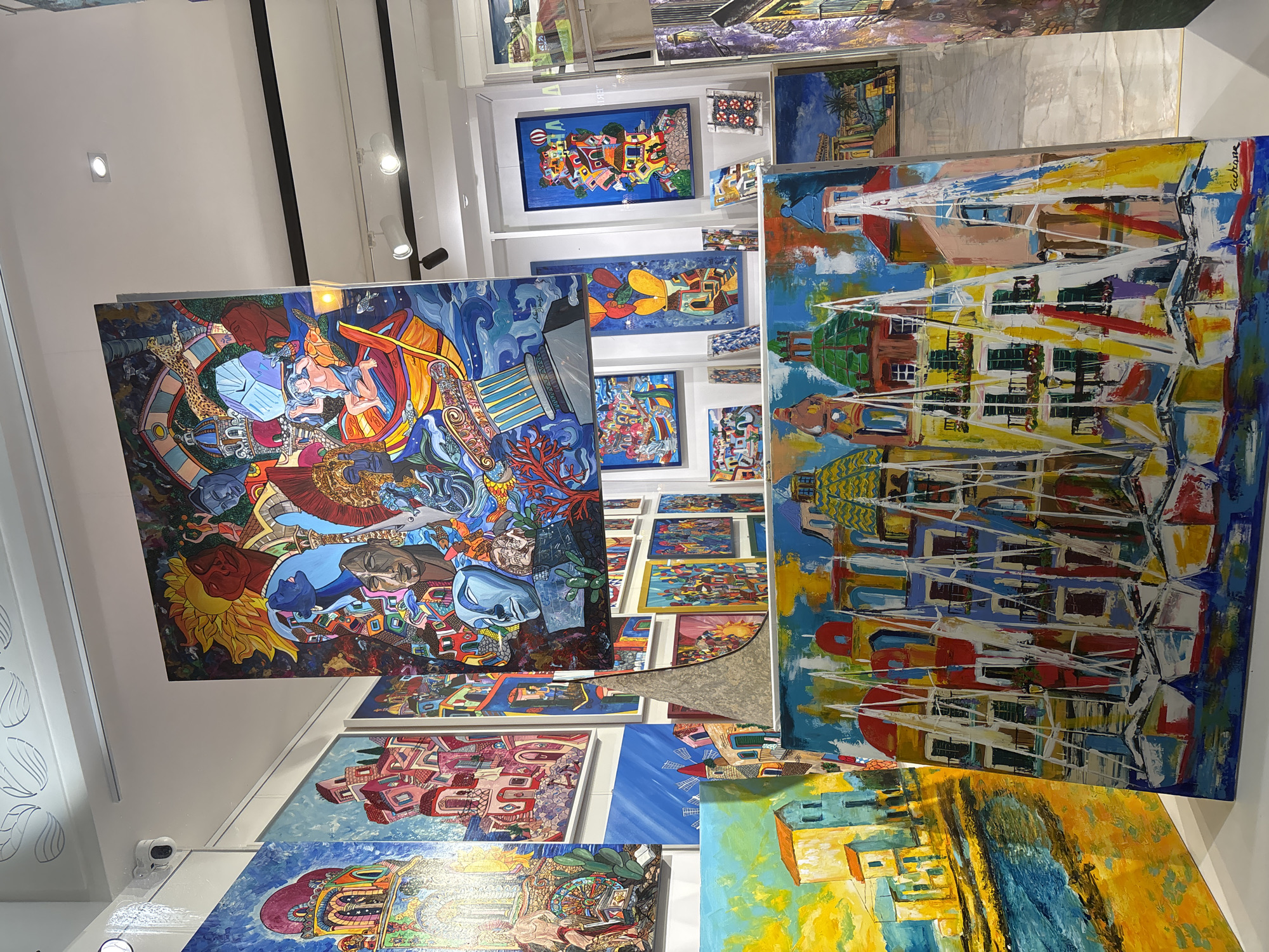 The image depicts an art gallery filled with vibrant and colorful paintings, possibly located in Palermo, Italy, given the style of architecture and maritime themes in some of the artwork. The gallery showcases a variety of paintings, many of which appear to be acrylic on canvas, with a distinct, expressive style characterized by bold brushstrokes and vivid colors.
In the foreground, a large painting features a colorful depiction of boats with white sails against a backdrop of buildings with red and green domed roofs. The style is reminiscent of Mediterranean coastal towns. To the left of this, another painting shows a sunlit scene of buildings along a body of water, with yellow and blue hues dominating.
Hanging from the ceiling, a large, complex painting is a focal point. It is a busy composition with many figures and symbolic elements, including a sun, a leopard, and various human faces and architectural structures rendered in a surreal and dynamic manner.
Other paintings on display include smaller works with similar architectural and landscape themes, some with a more abstract or patterned design. The gallery walls are painted white, and lighting fixtures are visible on the ceiling, suggesting a professional exhibition space. The overall impression is one of a lively and artistic environment.