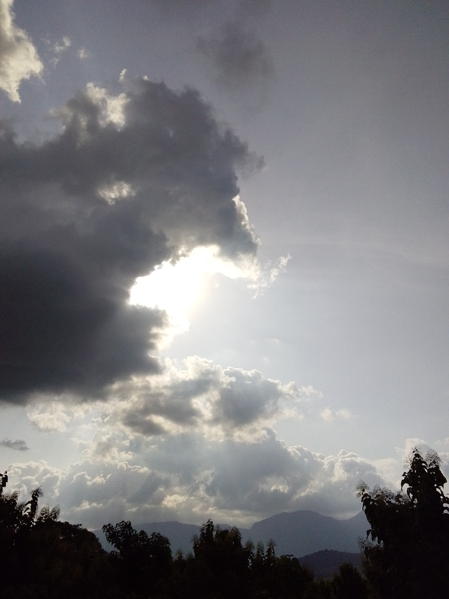 The image shows a dramatic sky over a landscape that is not in Serti, Nigeria. The sky is filled with dark, heavy storm clouds on the left, with a bright opening in the center where the sun is shining through, illuminating lighter clouds below. The right side of the sky is a pale, hazy grey. In the foreground, the silhouettes of trees and distant mountains are visible. The overall impression is of an impending or passing storm. The location is not identifiable as Serti, Nigeria based on the visual information presented.