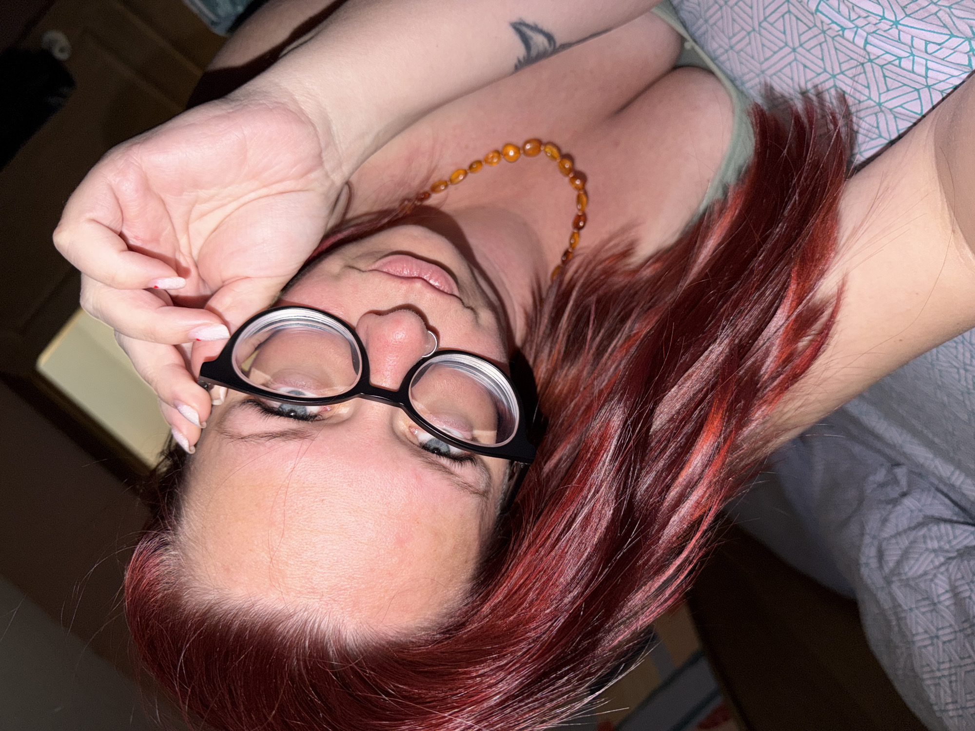 A woman with red hair is lying down and wearing glasses. She has a nose ring and a beaded necklace. She has a tattoo on her chest. The location is unknown.