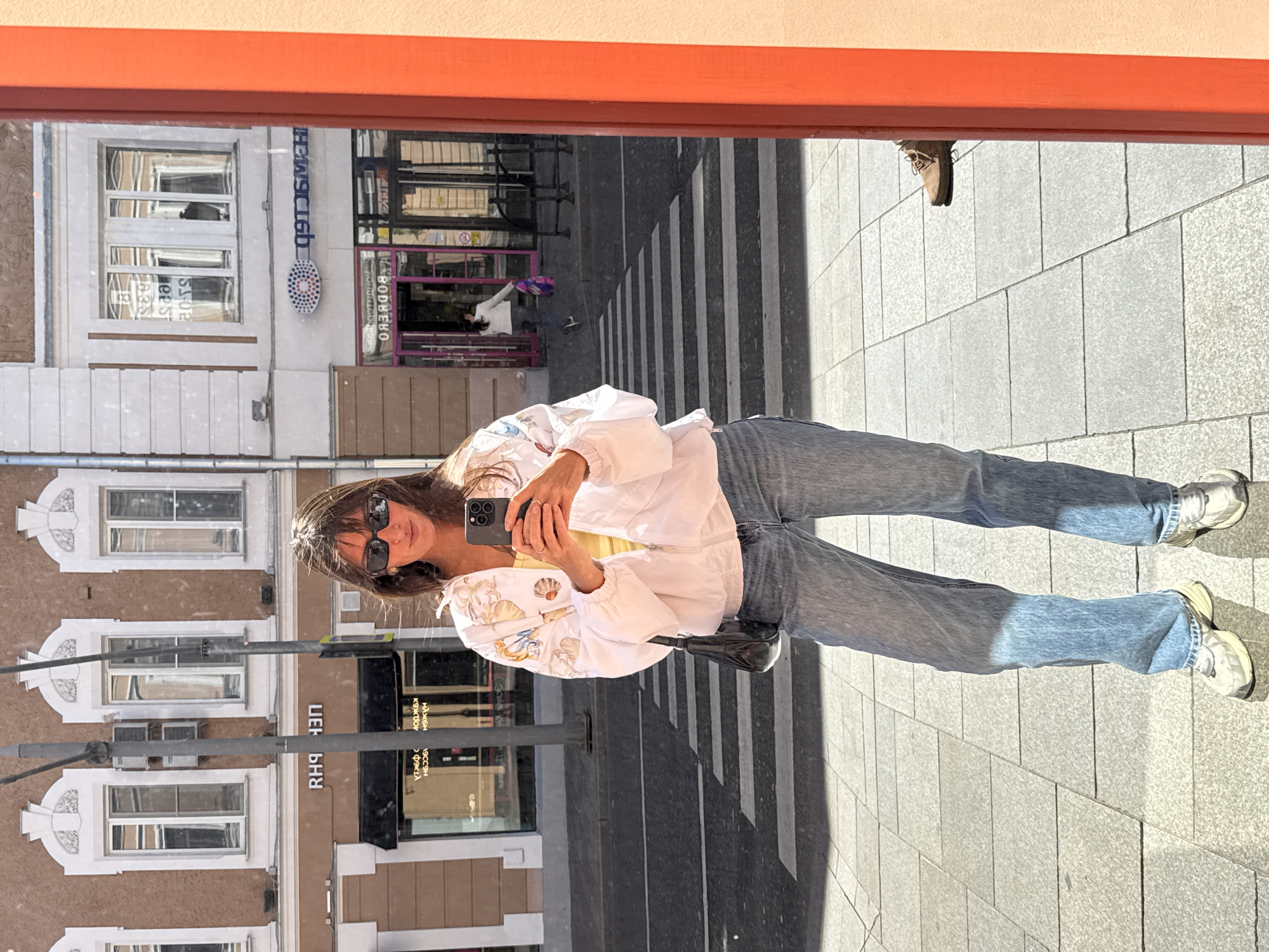 A young Caucasian woman with long dark hair and sunglasses is taking a selfie in a shop window. She is wearing a white jacket with a seashell print, a yellow top, and blue jeans. She is holding a black smartphone in her right hand. Her left hand is resting on her jacket pocket. She is standing on a paved sidewalk in front of a street with a crosswalk. In the reflection of the window, we can see buildings with shops. One of the shop signs has the text "RH9 43H" in Cyrillic letters. Another shop has a logo with blue and red circles. The building in the background has multiple windows with ornate detailing. A person in a white shirt and dark pants is walking across the street in the reflection. The environment appears to be a city street in Moscow, Russia, given the Cyrillic text on the signs.