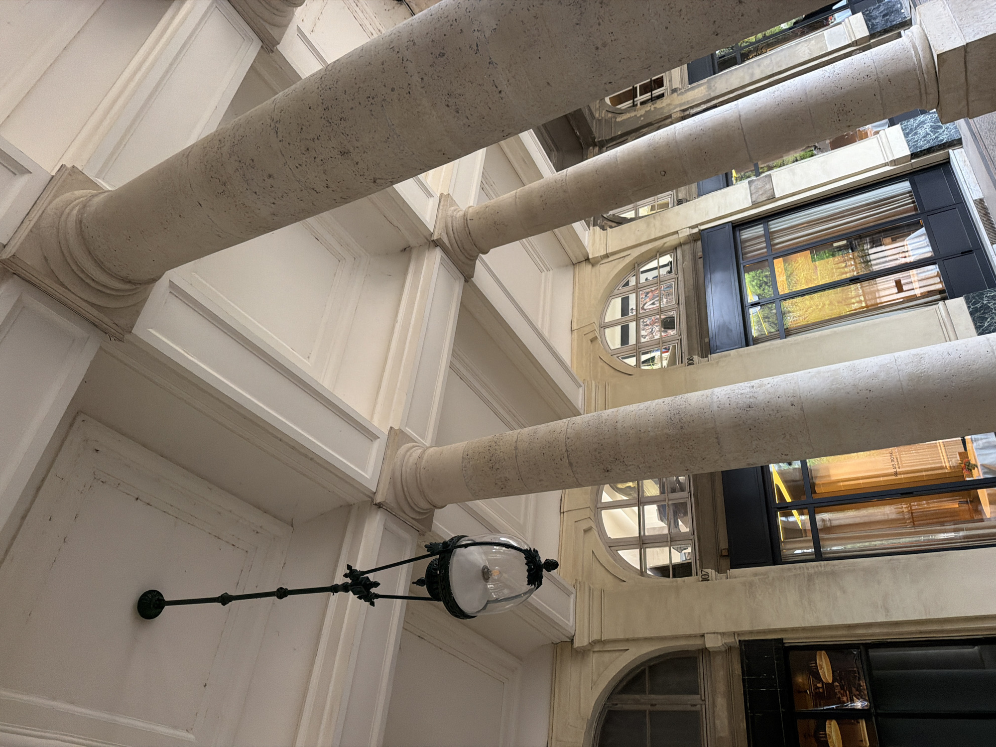 The image shows an upward-angled view of an interior architectural space. Several cylindrical columns run horizontally across the frame, their surfaces textured and colored in off-white or light beige. These columns appear to be supporting or embellishing a structure with multiple levels.
Between the columns, glimpses of an adjacent building are visible, with windows of various shapes (including circular and rectangular) and dark frames. Reflections in the windows suggest an outdoor environment.
In the lower-left quadrant, a vintage-style street lamp is mounted on a white wall. It features a clear glass globe and a dark-colored, ornate mounting arm.
The location is likely the Galerie Montpensier in the Palais Royal, Paris, France, based on the architectural elements and the overall ambiance of the scene. The image captures a blend of classical and contemporary architectural styles.