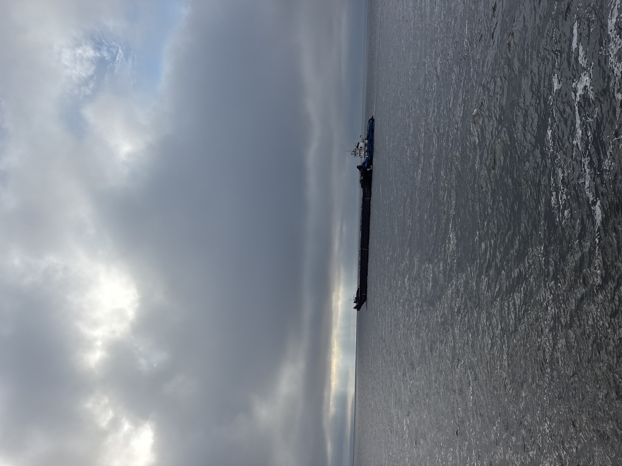 A barge, pushed by a tugboat, navigates a large body of water under a cloudy sky. Sunlight breaks through the clouds in visible rays, illuminating a portion of the water in the distance. The barge is long and dark, with a tugboat at its rear. The tugboat is blue and white, with a small flag visible on its cabin. The water is choppy, with whitecaps indicating moderate wind. The scene appears to be an overcast day in a port or waterway. The location is the city of Laishevo, Russia.