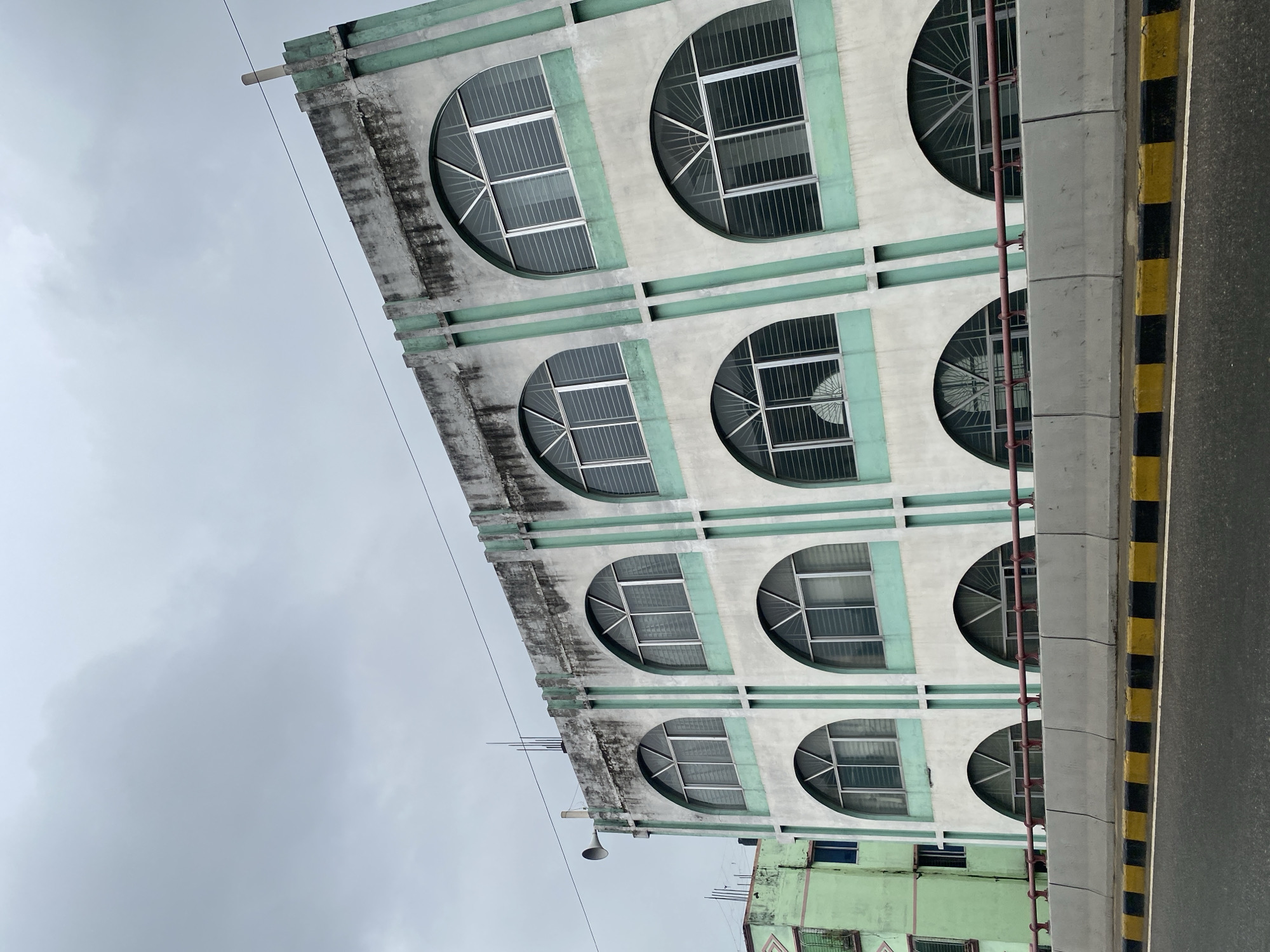 This image captures a portion of a multi-story building with a distinctive facade of arched windows, located in Dhaka, Bangladesh. The building's exterior is primarily white, accented with mint green vertical stripes and window frames. The windows themselves are large and arched, with white grilles covering their entirety. The building appears to be situated near a road, as evidenced by the presence of a concrete barrier and the asphalt surface of a roadway in the foreground. Yellow and black markings are visible along the edge of the road. The sky is overcast, with heavy gray clouds dominating the upper portion of the image. A smaller, green building is partially visible to the left of the main structure, suggesting a dense urban environment. A metallic speaker or horn is mounted on the side of the main building, near the roofline. The overall impression is of a commercial or institutional building in an urban setting with a somewhat dated architectural style.