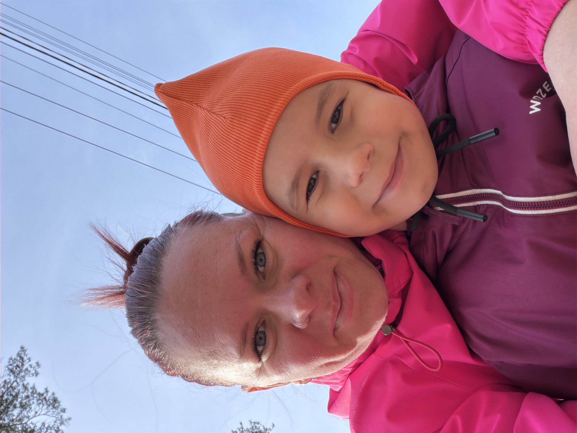 A selfie taken from below looking up at a woman and a young child, likely taken in Chaikovsky, Russia, is visible. The woman has her hair pulled up, fair skin and blue eyes. The child is wearing an orange knit cap and a maroon jacket with the word "WOZE" visible on the jacket. The woman is wearing a bright pink jacket. Power lines are visible in the top left corner of the image with a pale blue sky in the background. Bare trees are visible in the bottom left corner of the image.
