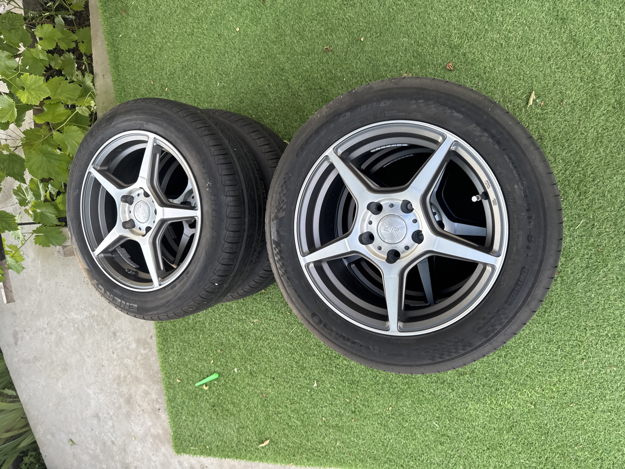 Four alloy wheels with tires are stacked on artificial turf. The top wheel is partially obscured by the wheel below it. A small green object is on the concrete in the upper left portion of the image. The wheels are silver and have a five-spoke design. The tires appear to be brand new, with no signs of wear. The background shows a concrete walkway and some green foliage. The scene is outdoors, with natural lighting. The location is specified as Dobrovske settlement, Ukraine.