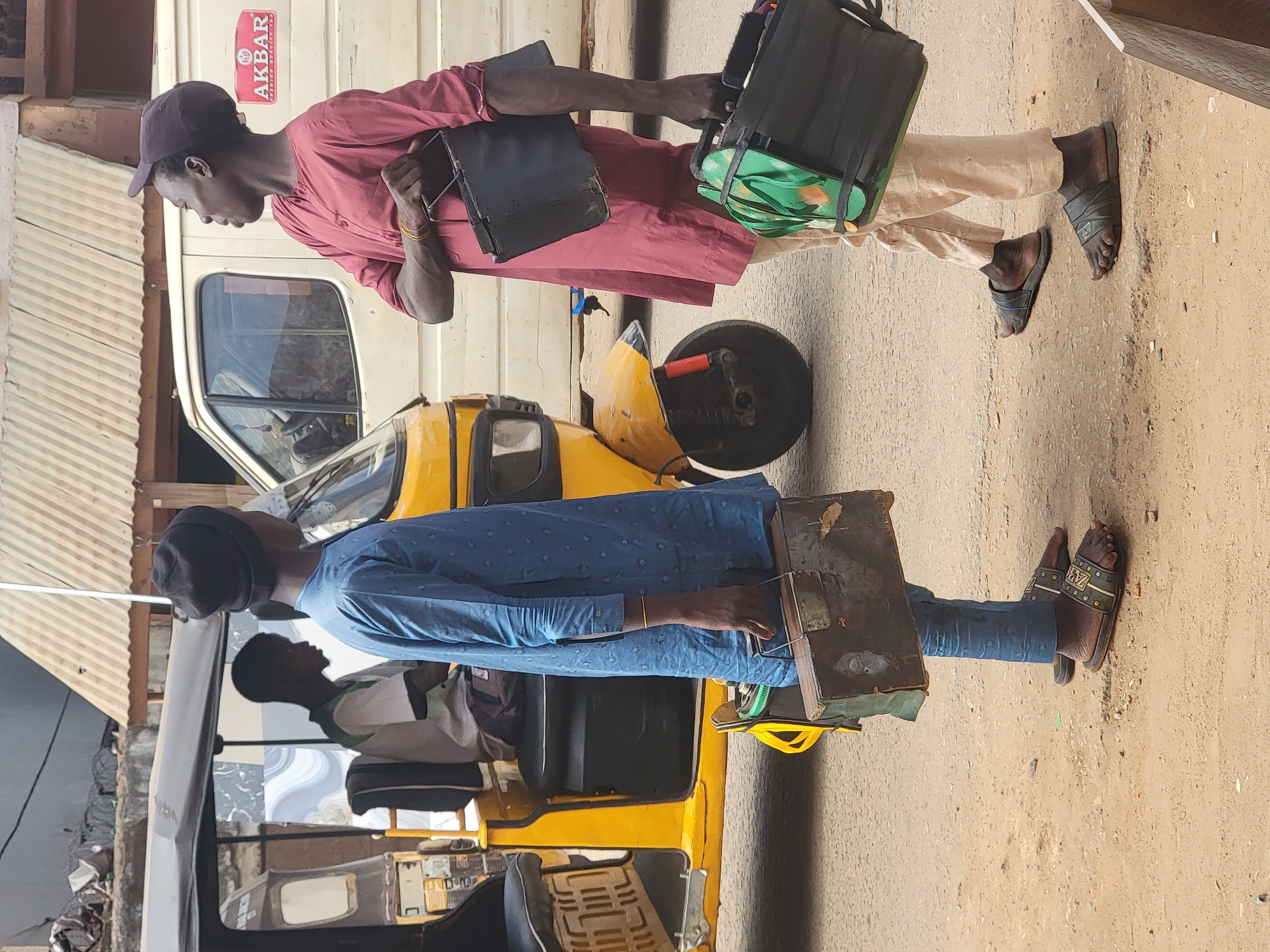 The image shows two men standing in what appears to be Maiburiji, Nigeria. The man on the right wears a maroon shirt, khaki pants, a baseball cap, and sandals. He carries a black bag and a flat, dark object, possibly a briefcase or folder. The man on the left wears a blue shirt and pants, a kufi cap, and sandals with branding on the straps. He also carries a box-like object. Both men stand on a tan-colored surface beside a yellow, three-wheeled vehicle. The background includes buildings with corrugated metal siding. A vehicle bearing the name "AKBAR" is visible.