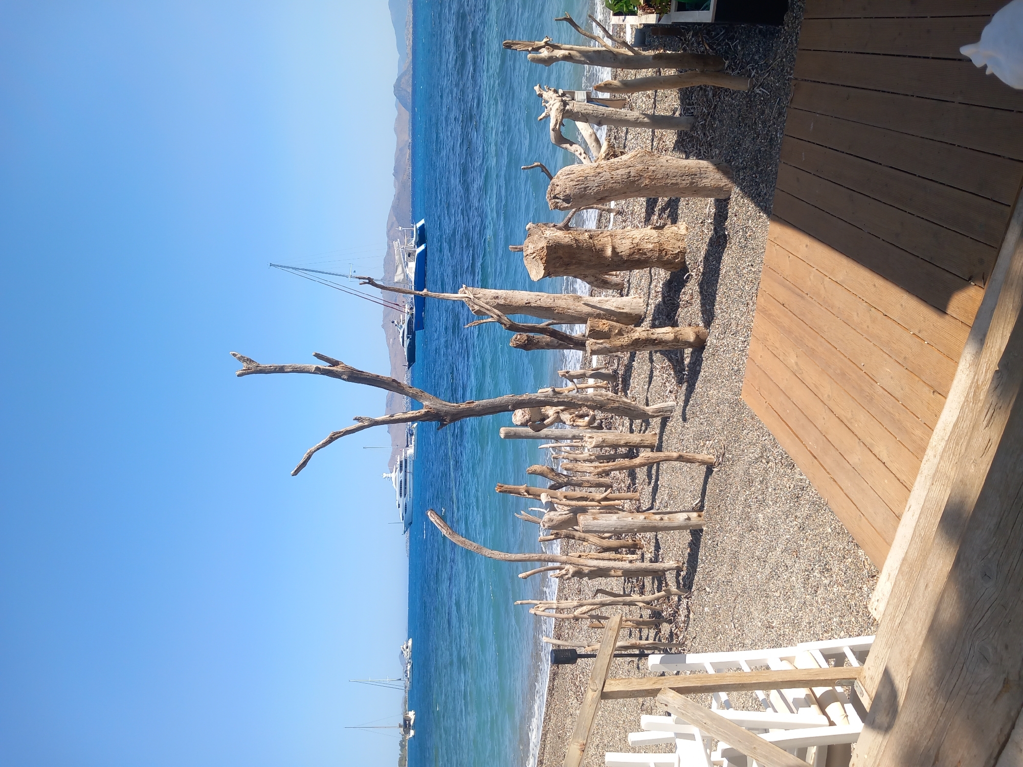 The image shows a waterfront scene in Kos, Greece. In the foreground, a wooden deck with planks is visible, partially in shadow. Beyond the deck is a pebble beach adorned with numerous pieces of driftwood of varying sizes, arranged upright and horizontally, resembling a rustic sculpture garden. In the background, the turquoise Aegean Sea stretches out under a clear blue sky. Several boats and yachts are anchored or sailing on the water, including a large white yacht and two sailboats. The coastline in the distance features rolling hills. White outdoor furniture is partially visible on the left side of the image. The overall impression is a serene, seaside setting with artistic elements.
