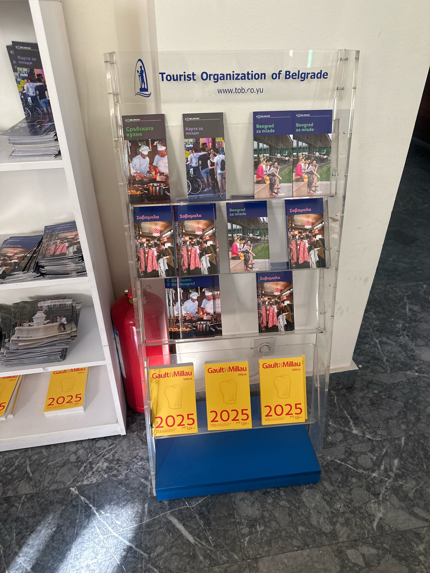 The image depicts a tourist information display stand in Belgrade, Serbia. The stand is made of clear acrylic and is filled with brochures. Prominently displayed at the top is the logo and text "Tourist Organization of Belgrade" along with the website "www.tob.co.yu". Below this, there are multiple shelves holding various brochures. Some brochures have Cyrillic text and images of food preparation, suggesting they are related to Serbian cuisine. Others have images of young people and text indicating "Beograd za mlade" (Belgrade for youth). Further down, there are yellow brochures with "Gault&Millau SRBIJA 2025" printed on them. A bright red fire extinguisher is visible to the left of the display stand. The flooring appears to be dark marble. The lighting suggests an indoor environment, possibly a public building or tourist center.