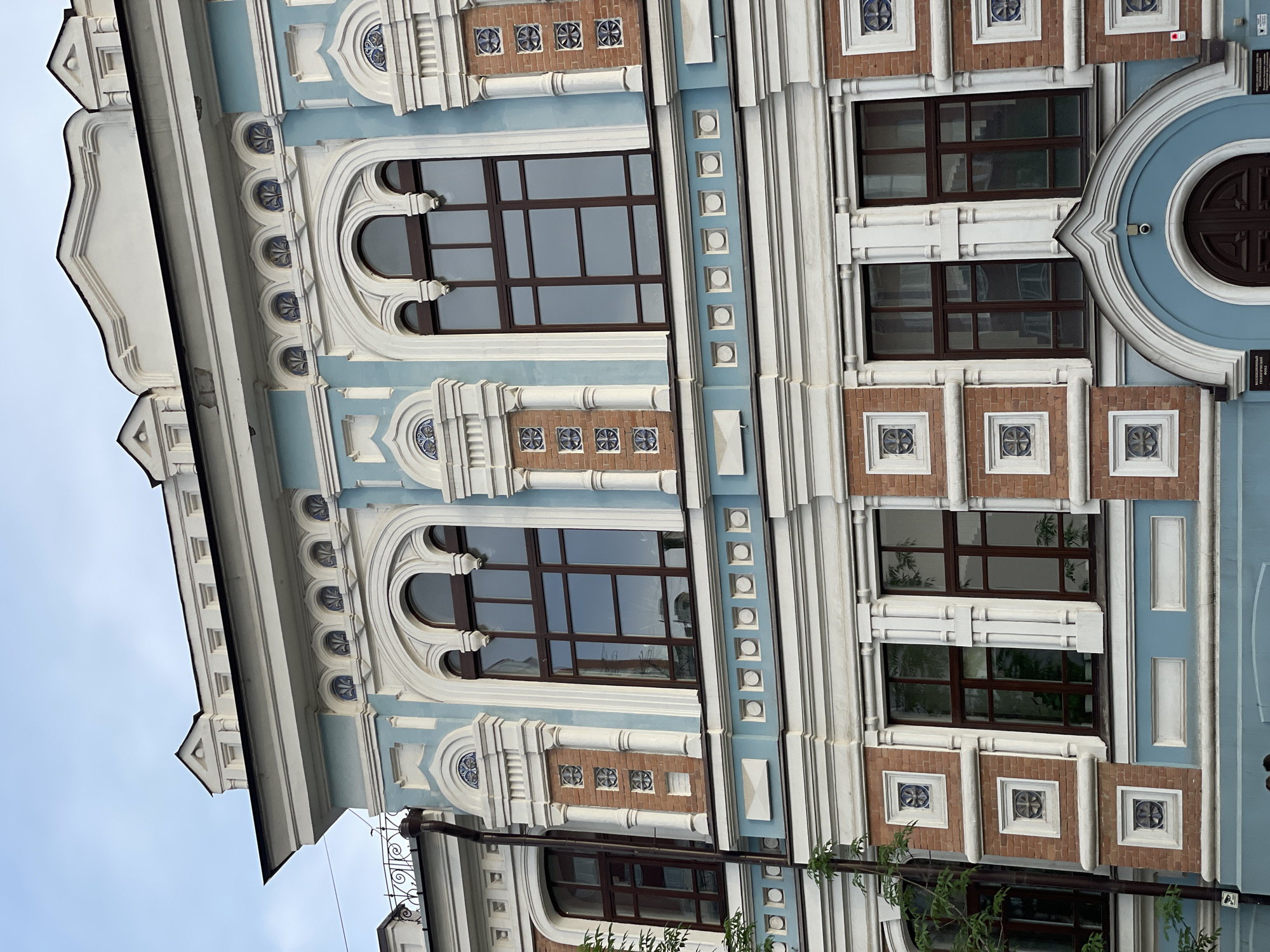 The image shows the facade of a building with ornate architecture. The building has a light blue and white color scheme with brick accents. There are several large windows with dark frames. The windows on the upper level are arched, while those on the lower level are rectangular. Decorative elements, such as pilasters and carved panels, adorn the facade. The sky in the background is overcast. On the lower part of the building, there appears to be an entrance with a dark wooden door. There are also signs with text in Russian on the lower portion of the building, suggesting this is a public or official building. Based on the architectural style and the text on the signs, this building is likely the "Krasnoyarsk State Pedagogical University named after V.P. Astafyev" located in Krasnoyarsk, Russia. There are no people visible in the image.