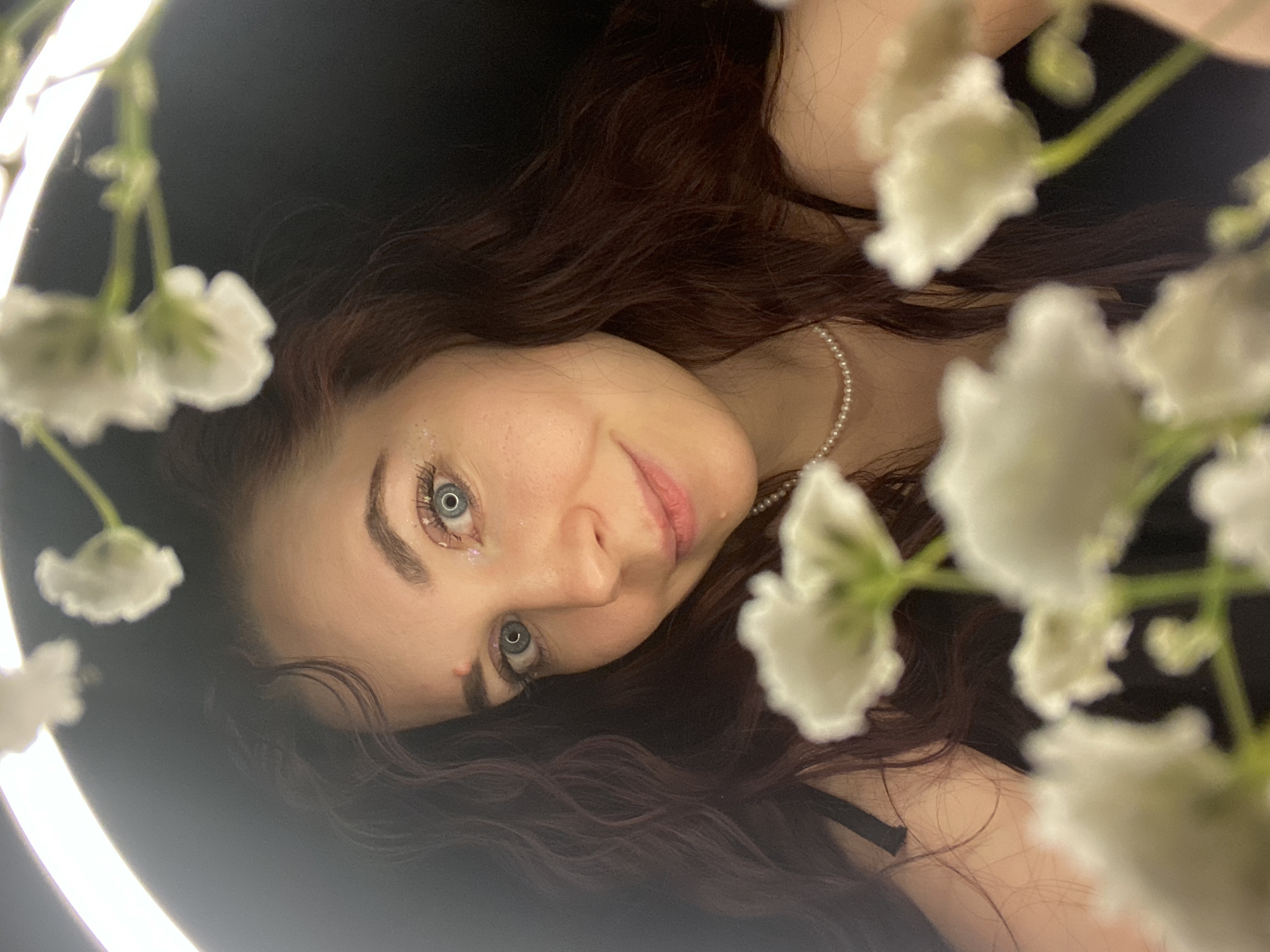 A close-up photograph shows a woman with pale skin, reddish-brown hair, and light blue eyes. She is tilted at an angle, looking directly at the camera. Her eyes are decorated with small, sparkly star-shaped stickers. A delicate pearl necklace is visible around her neck. Her hair is loose and wavy, surrounding her face. Sprigs of small white flowers are in the foreground, partially obscuring her face and body. A ring light is visible as a bright halo surrounding the scene. It is impossible to verify the location is in Chaikovsky, Russia with the information provided.